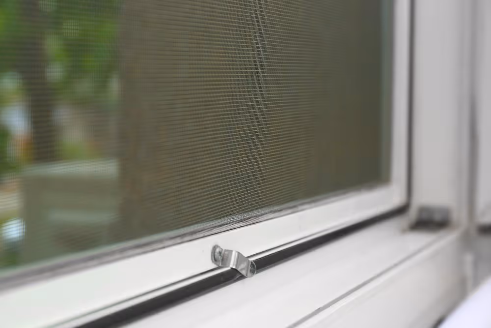 Detail okenního rámu z bílé PVC s kovovou sponou a zeleným pozadím venku.