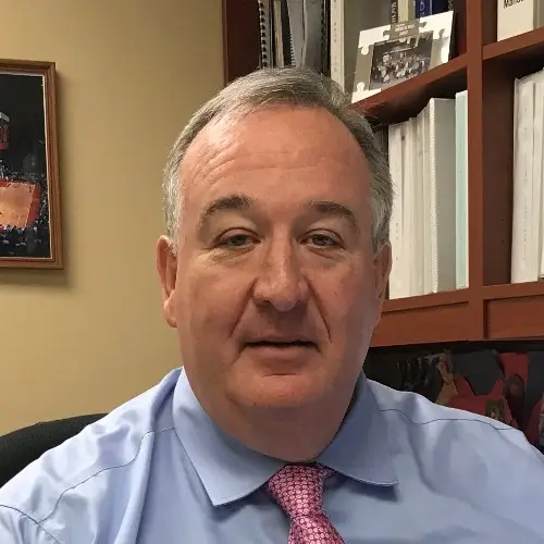 Man in blue dress shirt and pink tie sitting at office desk with bookshelf, representing AJTC IT support leadership