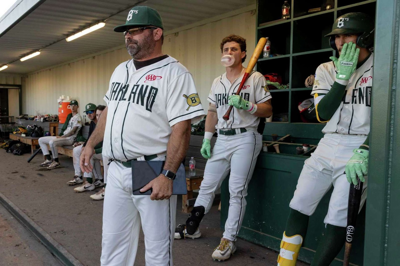A view of the Oakland Baller’s dugout. One player blows a pink bubble gum bubble and holds a bat. The manager stands at the front, tablet in hand. The Oakland Ballers became the first AI-managed professional sports team, underscoring the growing role of AI in baseball.
