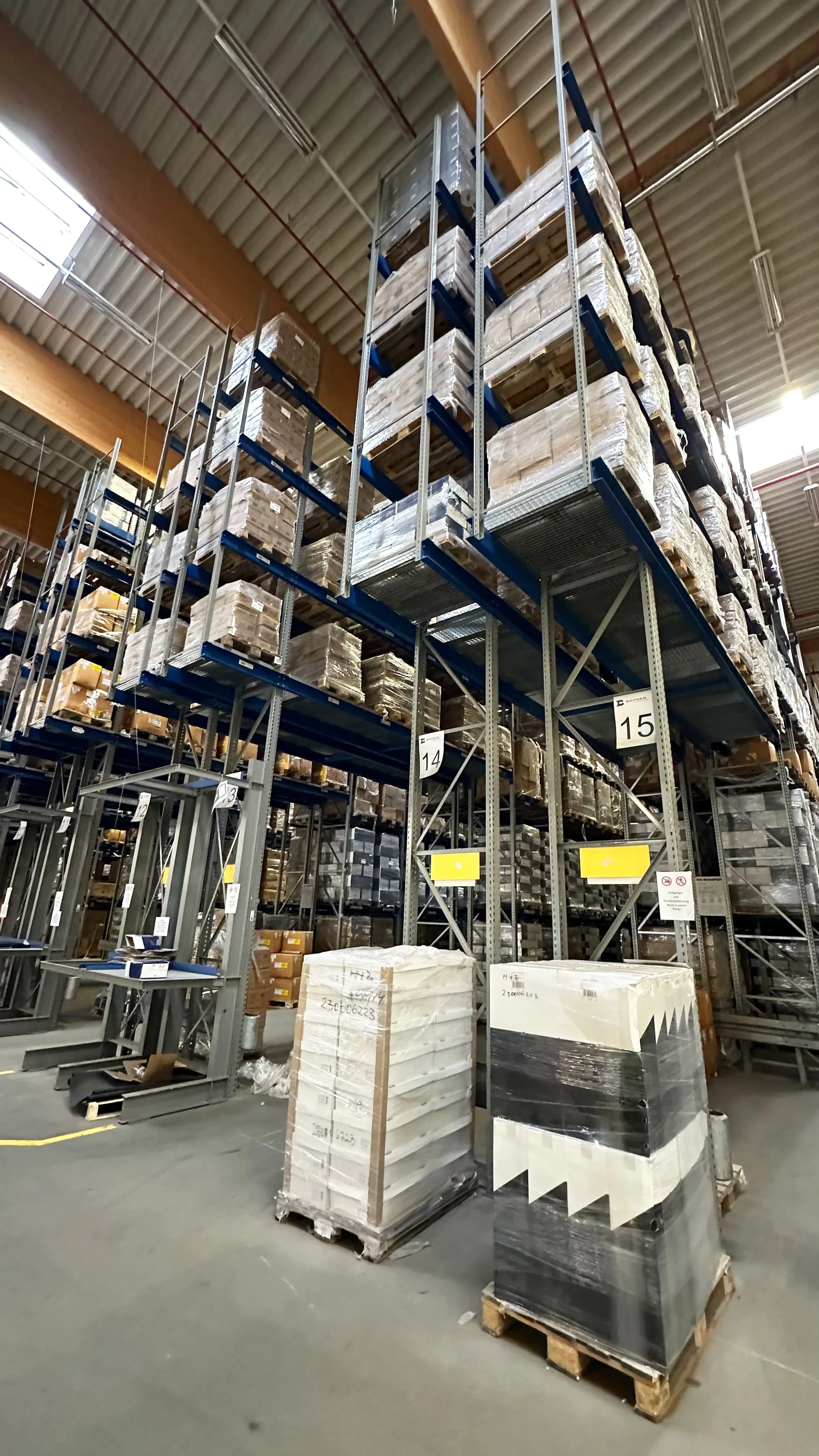 Lagerhalle mit hohen Metallregalen, die mit Paletten voller verpackter Waren bestückt sind, sowie zwei Stapeln verpackter Paletten auf dem Betonboden.