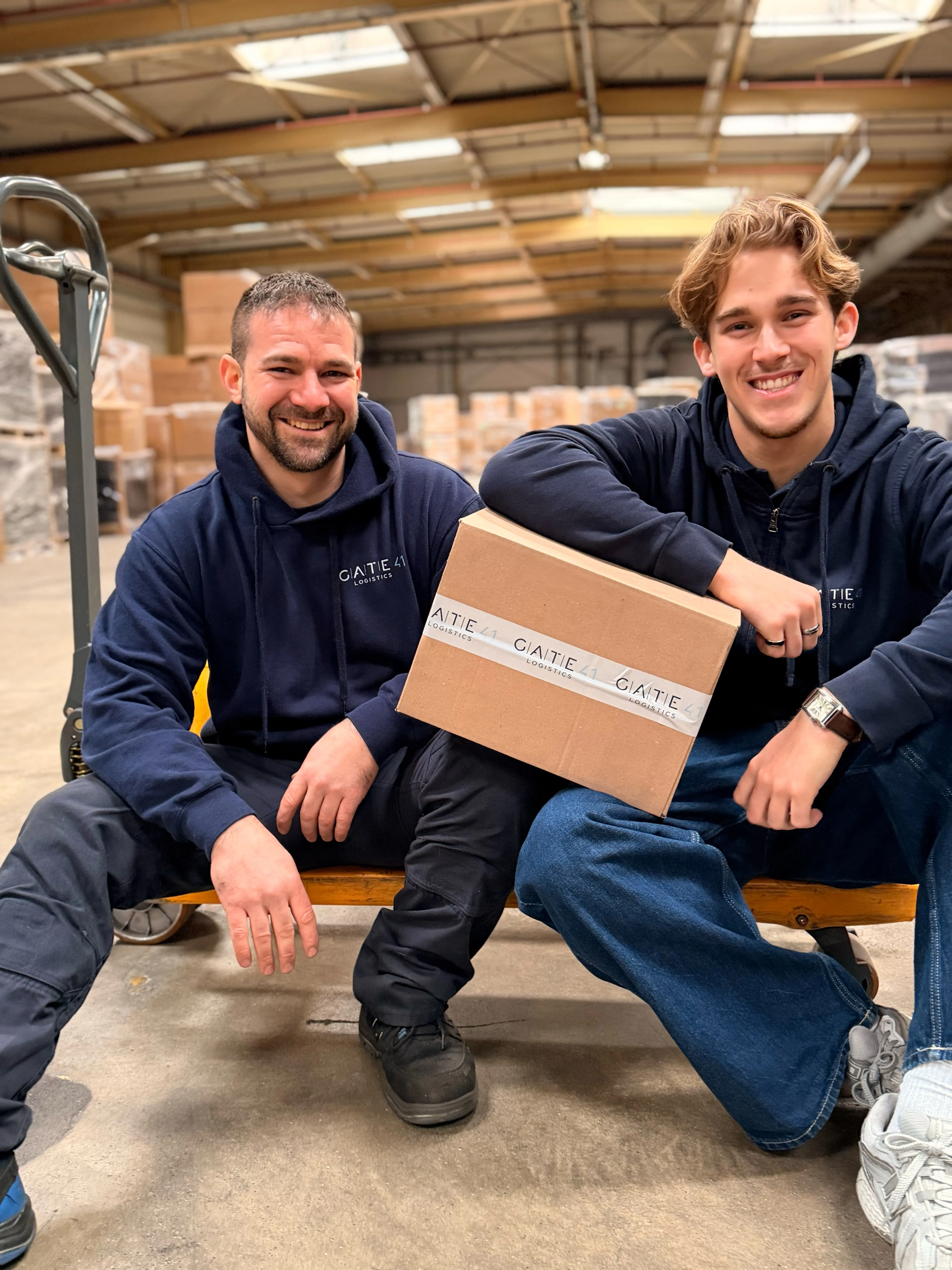 Zwei lächelnde Männer in dunkelblauen Kapuzenpullis, die in einem Lagerhaus eine Kiste mit der Aufschrift „GATE 41 LOGISTICS“ in den Händen halten.