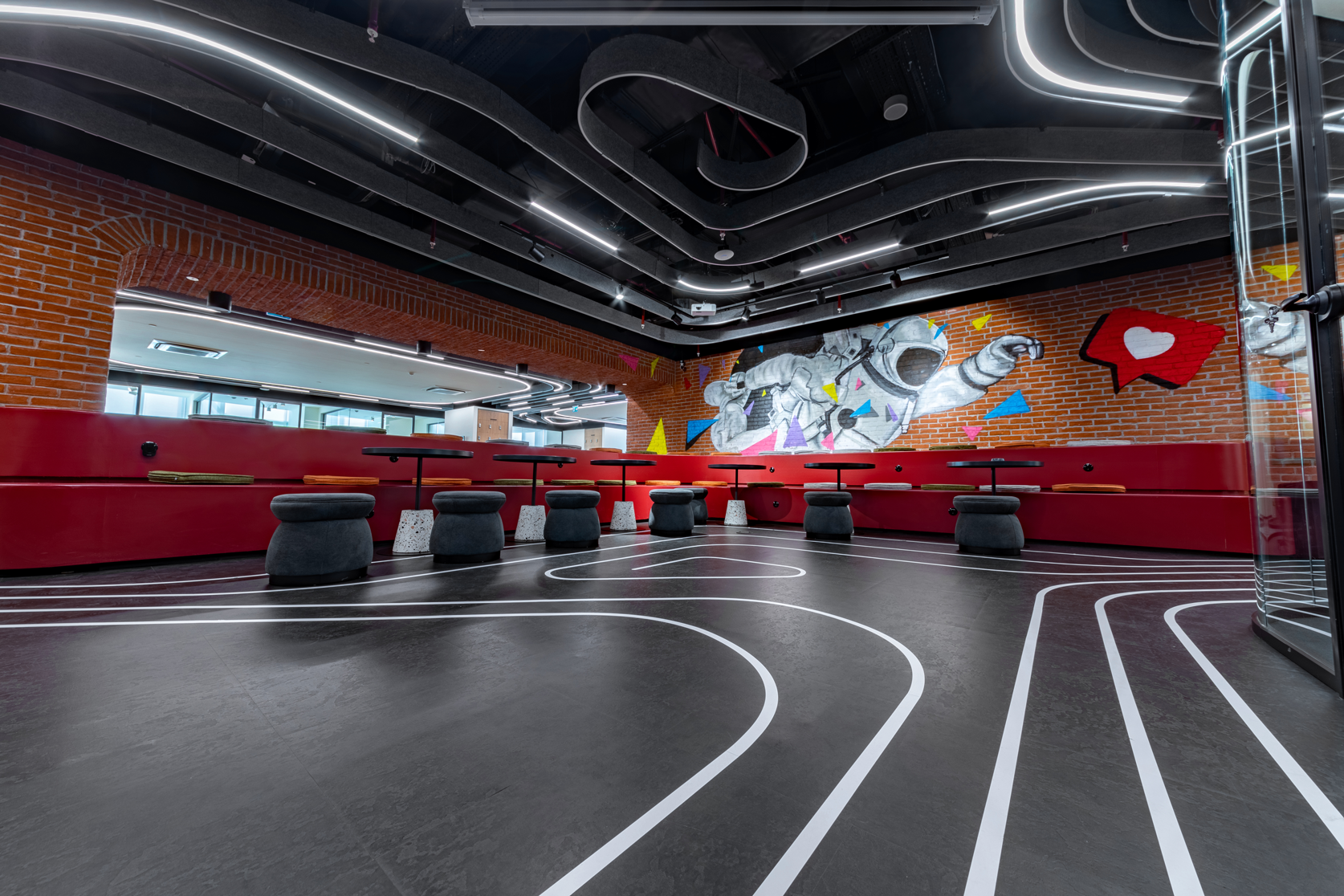 Tooplay office lounge area featuring a brick wall with astronaut graffiti, red bench seating, and dynamic floor markings resembling a race track.