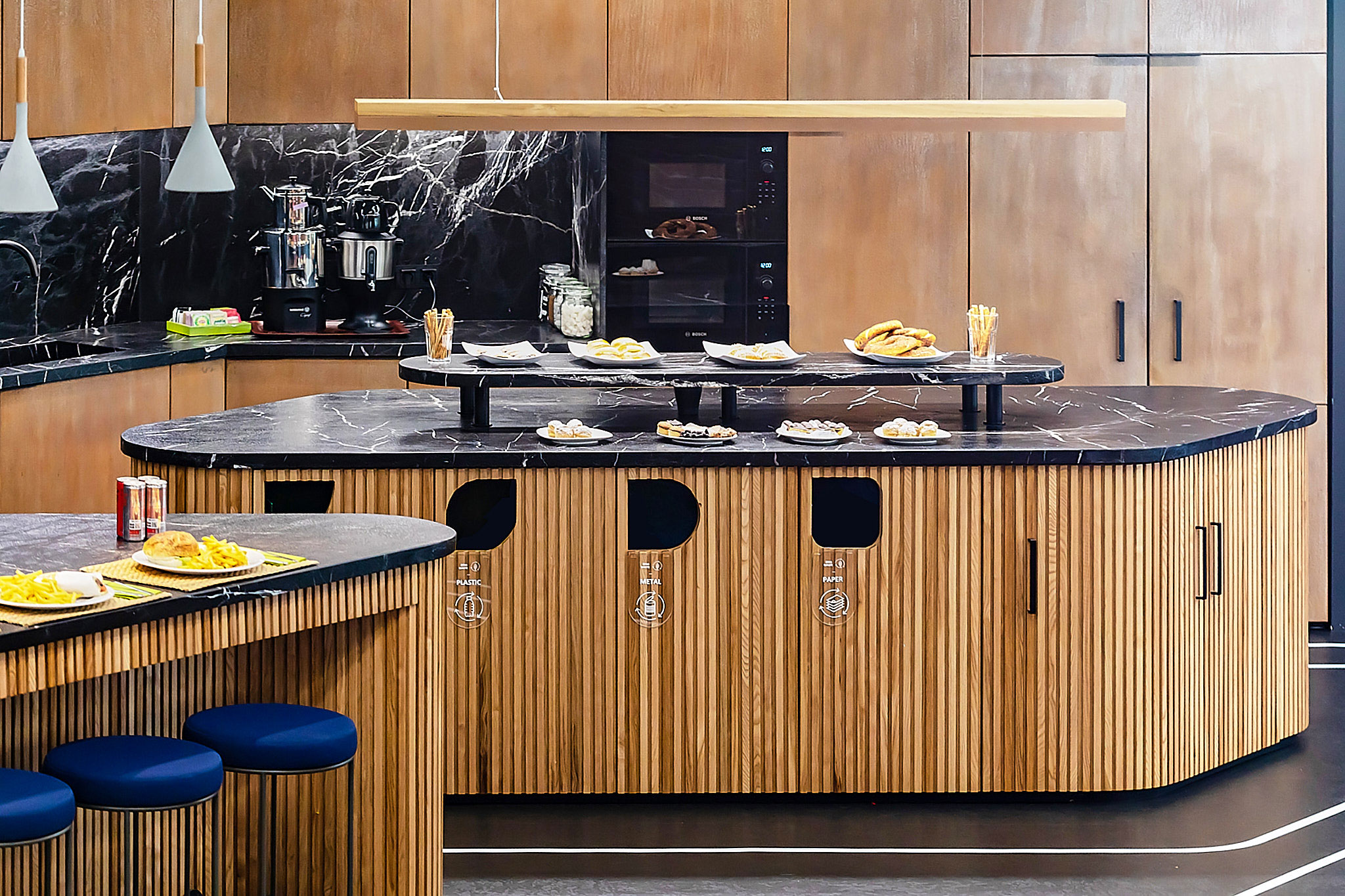 Modern office kitchenette with marble countertops, wooden slat islands, and integrated recycling stations for sustainability.