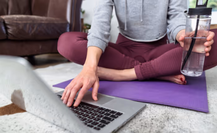 a home living room with a white woman, sitting down on a purple yoga mat, wearing workout clothes, holding a bottle of water and reaching for her laptop.