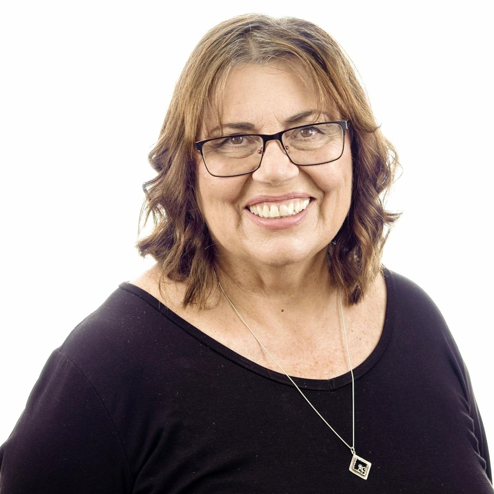 Smiling middle-aged woman with glasses, wavy shoulder-length brown hair, black top, and a silver necklace with a square pendant.