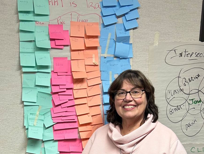 Smiling woman with glasses and dark hair in a light pink sweater stands in front of a wall covered with colorful sticky notes and handwritten charts.