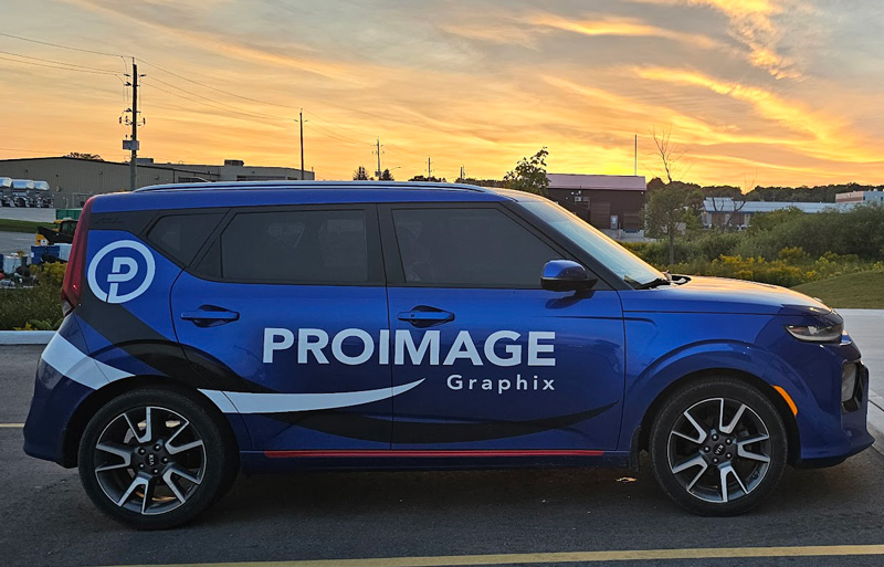 A blue car with "PROIMAGE Graphix" and a logo on the side is parked in a lot. The background features industrial buildings and a vibrant sunset sky.