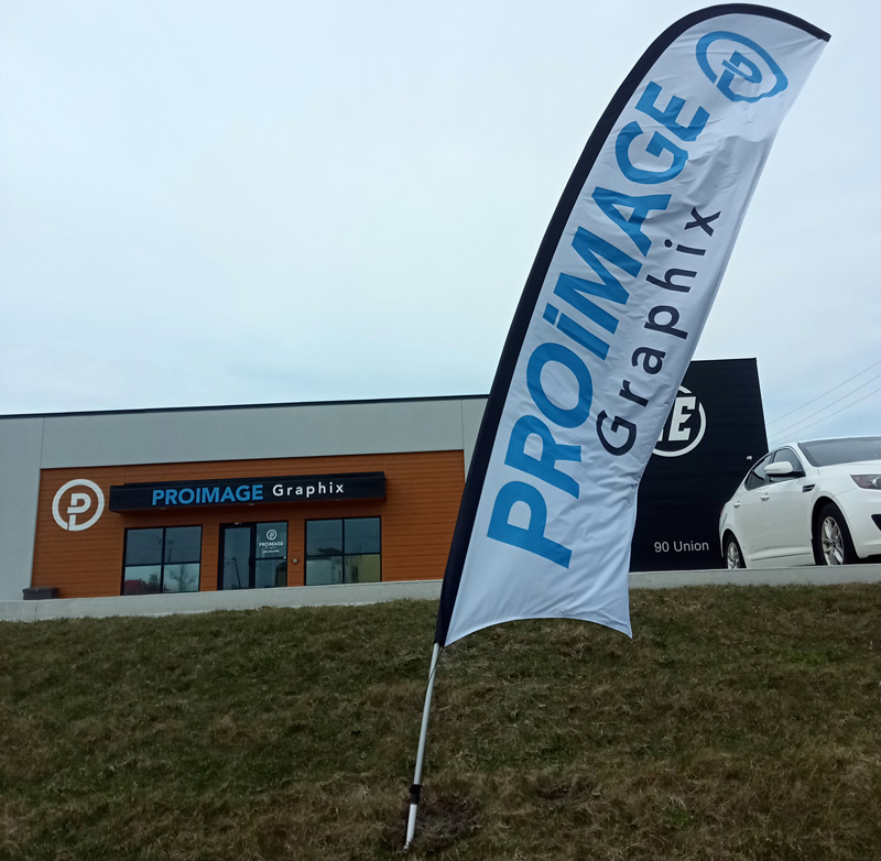 A standing banner displays "PROIMAGE Graphix" on a grassy hill. In the background, a building has a sign with the same name. A white car is parked nearby.