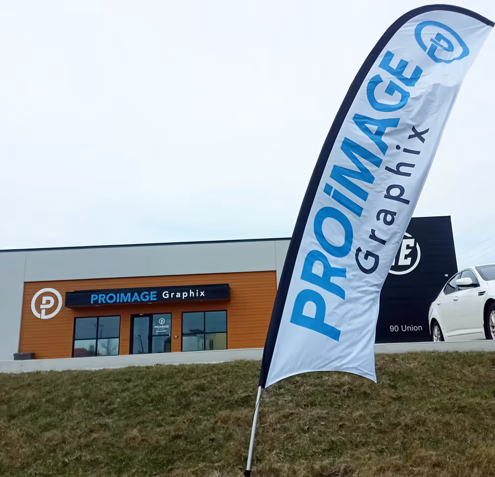 A standing banner displays "PROIMAGE Graphix" on a grassy hill. In the background, a building has a sign with the same name. A white car is parked nearby.