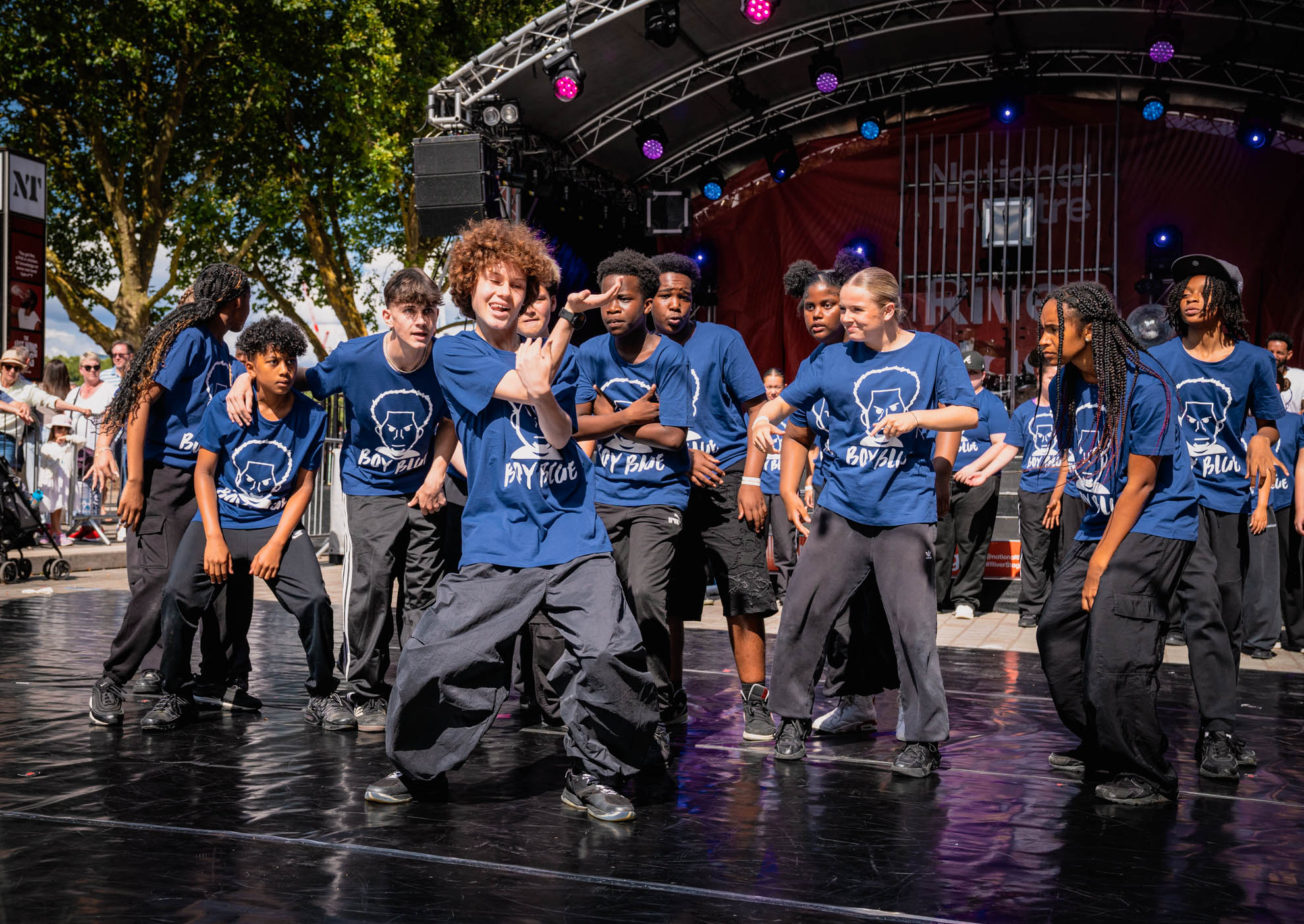 Group of young dancers performing on an outdoor stage wearing matching blue 'Boy Blue' t-shirts and black pants under a canopy with colorful lights.