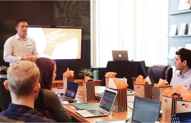 Man standing and speaking in front of a screen presenting to a group of seated people around a conference table with laptops and gift bags.