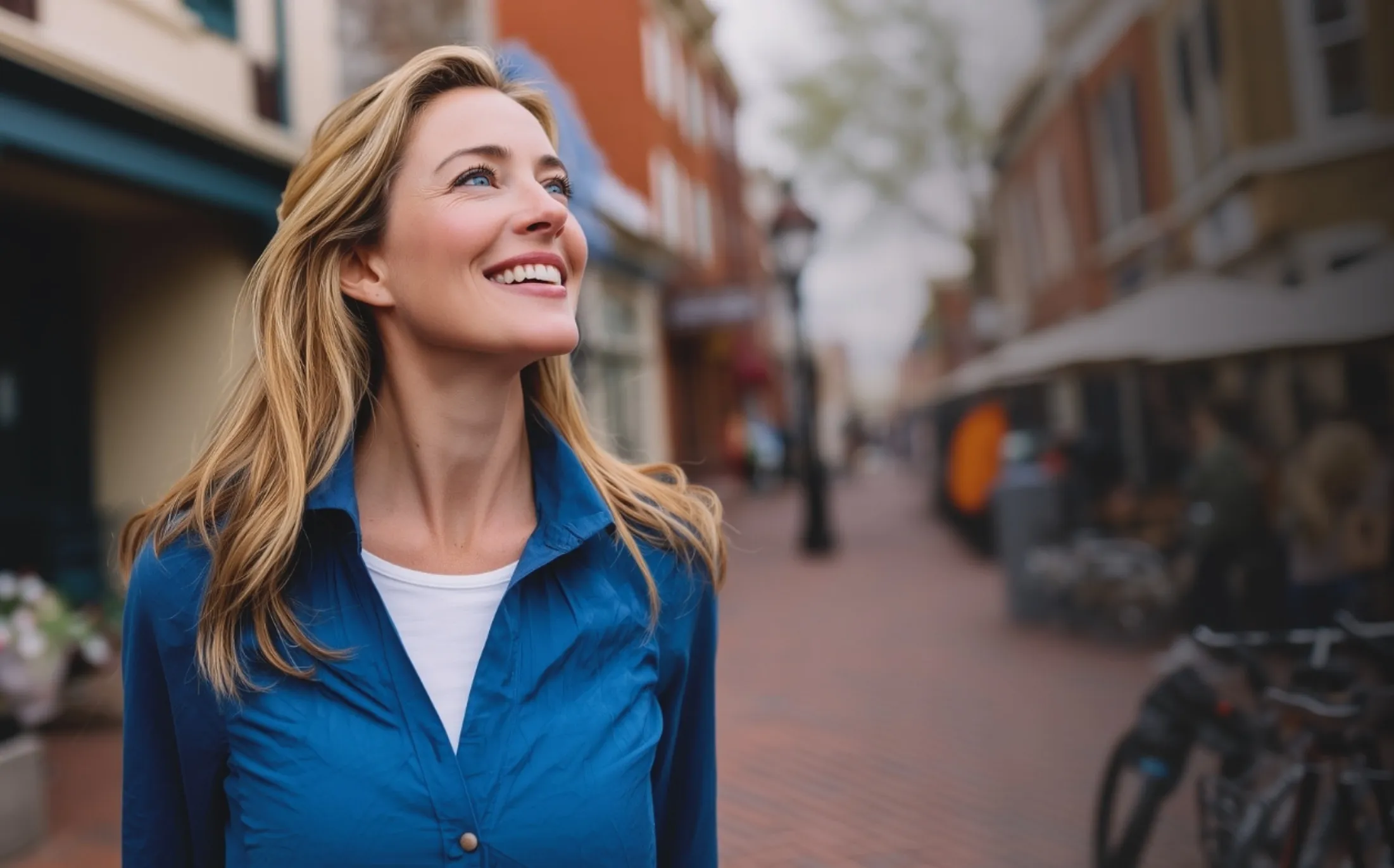Smiling woman with blonde hair wearing a blue shirt walking on a brick-paved street in an urban area.