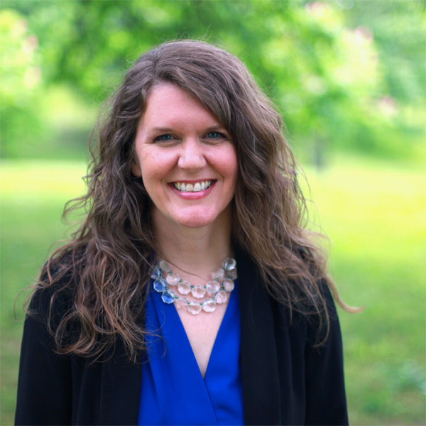 This image is of Kerri Johannsen, a white woman with long dark wavy hair. She's wearing a royal blue blouse with a black jacket and a necklace of clear beads. Kerri is smiling as she stands outside, surrounded by green trees and grass.