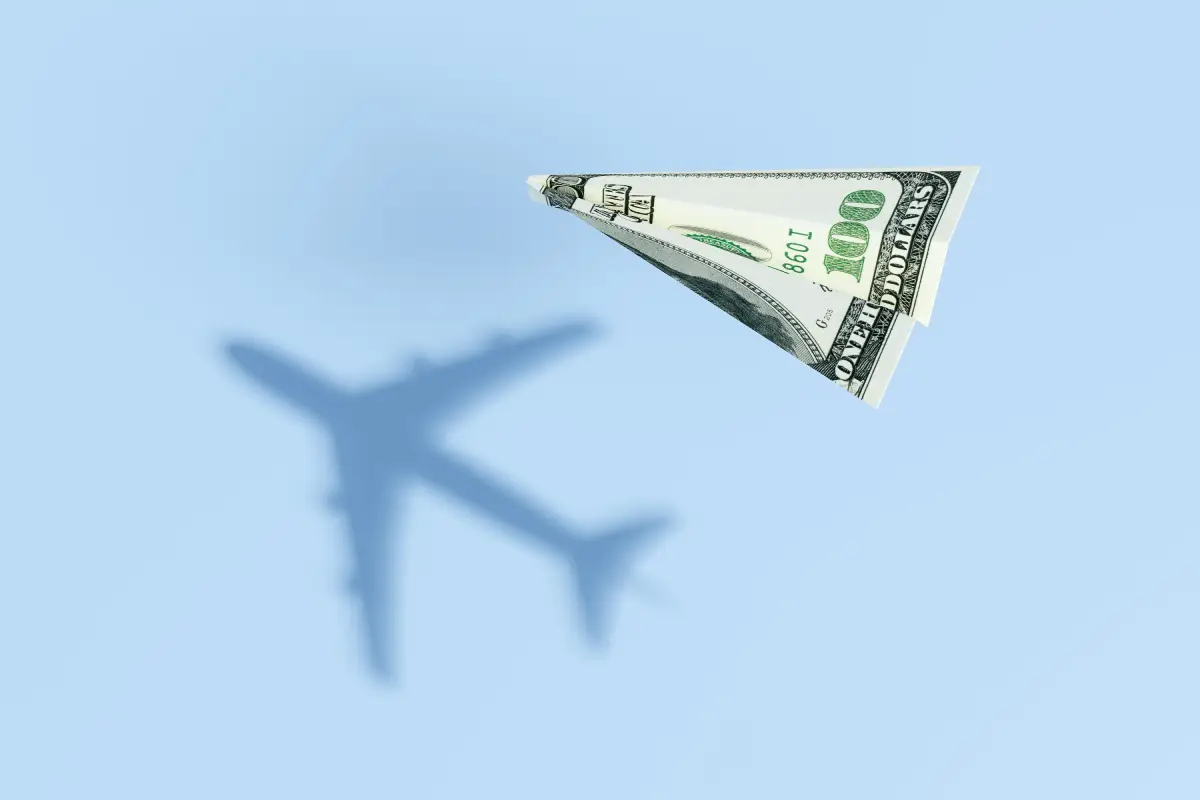 100-Dollar-Schein als Papierflieger. Im Hintergrund sind als Schatten Umrisse eines echten Flugzeugs zu sehen.
