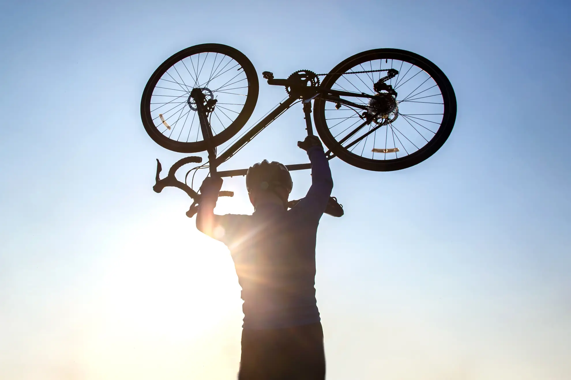 Radfahrer streckt erfreut sein Rennrad in die Luft. Im Hintergrund sind blauer Himmel und ein paar Sonnenstrahlen zu sehen.