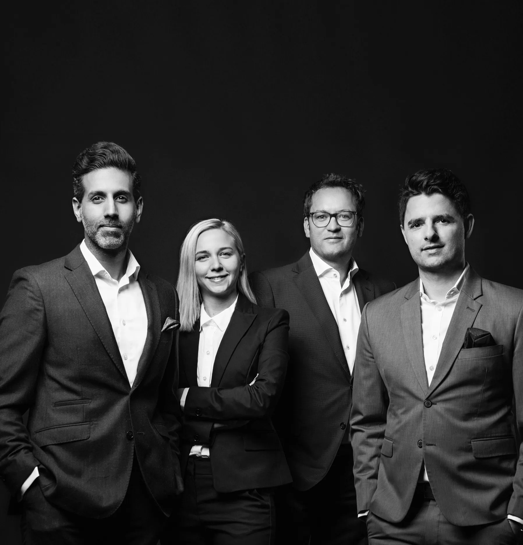 Black and white portrait of four professionally dressed adults standing in a row against a dark background, including three men in suits and a woman in a suit with arms crossed, all facing the camera with confident expressions.