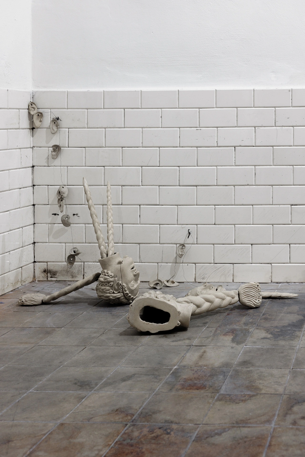 Anzhelika Palyvoda, ceramic sculptural cluster on tiled floor with braided horns, hollow heads, and wall-hung ceramic fragments, 2026