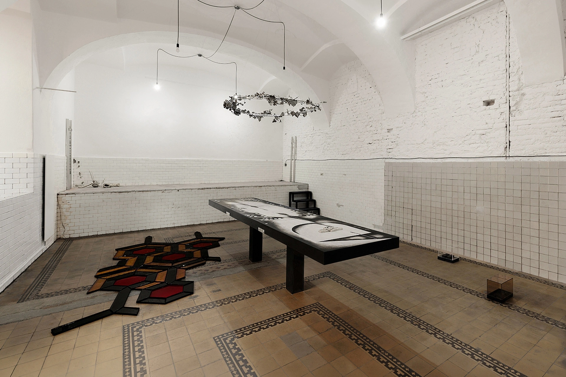 Installation overview, Echoes That Refuse to Fade, FAVU Gallery Brno, white-tiled hall with black table, wreath suspension, and hexagonal grid, 2026