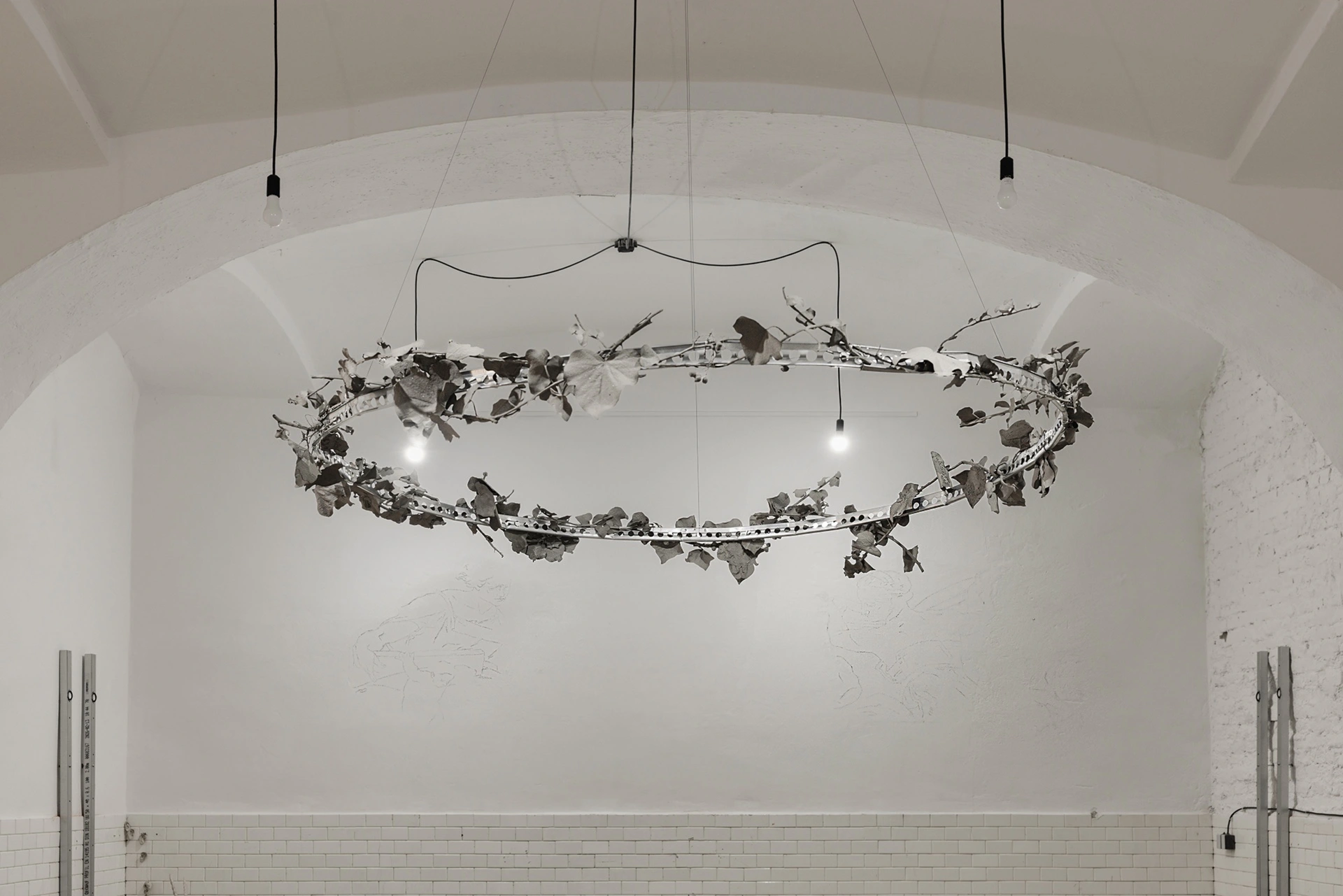 Céline Struger, wreath chandelier of steel ring and dried grey-black leaves with bare filament bulbs, suspended in vaulted hall, 2026