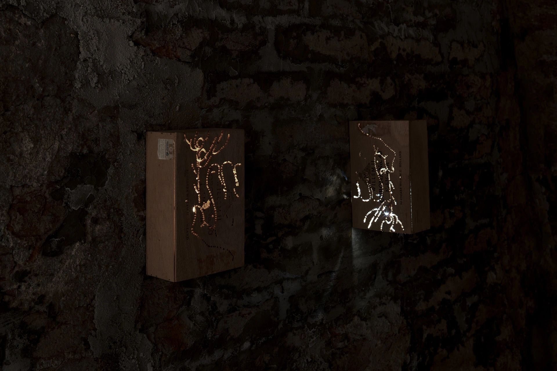 Anzhelika Palyvoda, two backlit cardboard boxes with pierced figurative silhouettes casting shadow onto rough brick wall, 2026