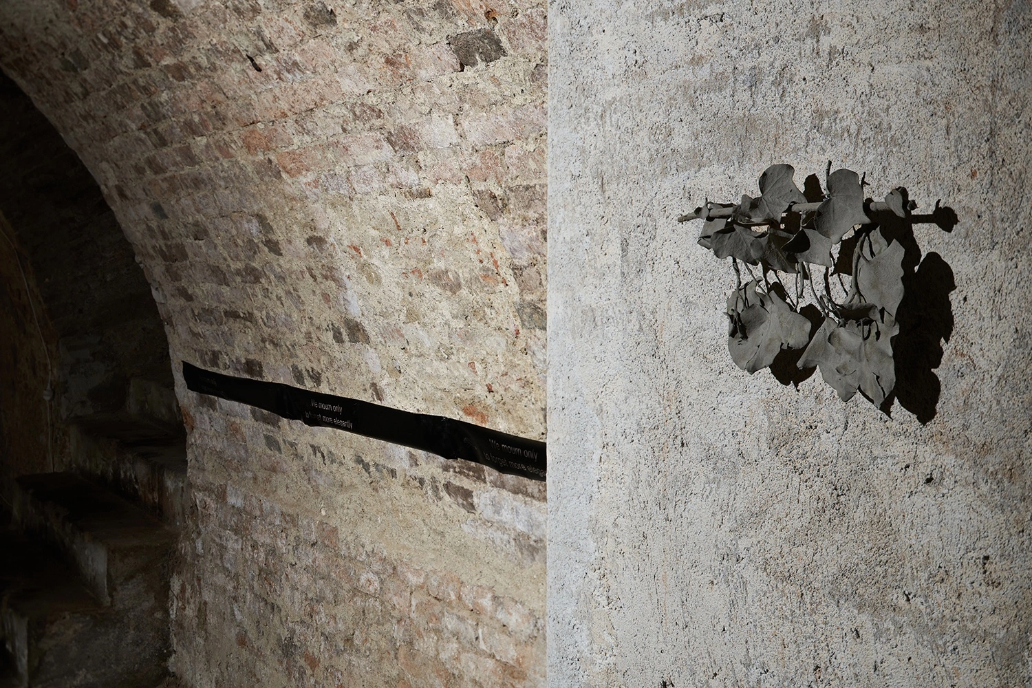 Anzhelika Palyvoda, brick wall detail with exhibition label and ceramic leaf cluster mounted on arch, underground passage, 2026