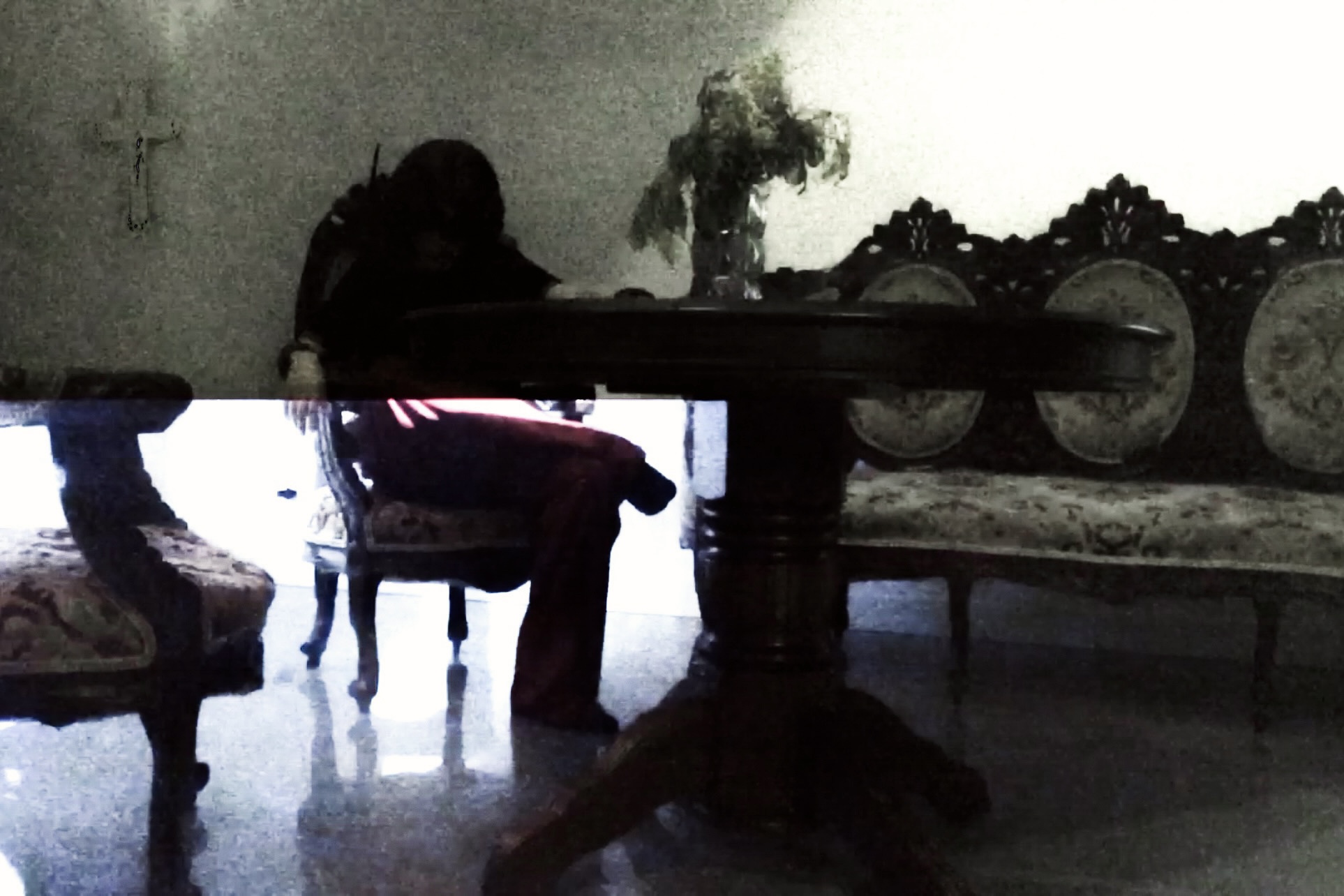 Spread from TheHunterIssue Vol.1 by Leo Shmunk, grainy domestic interior with a hooded figure slumped at a polished wooden table, dried bouquet and small wall crucifix behind, ornate carved settee at right, 2026. Photo: Leo Shmunk.