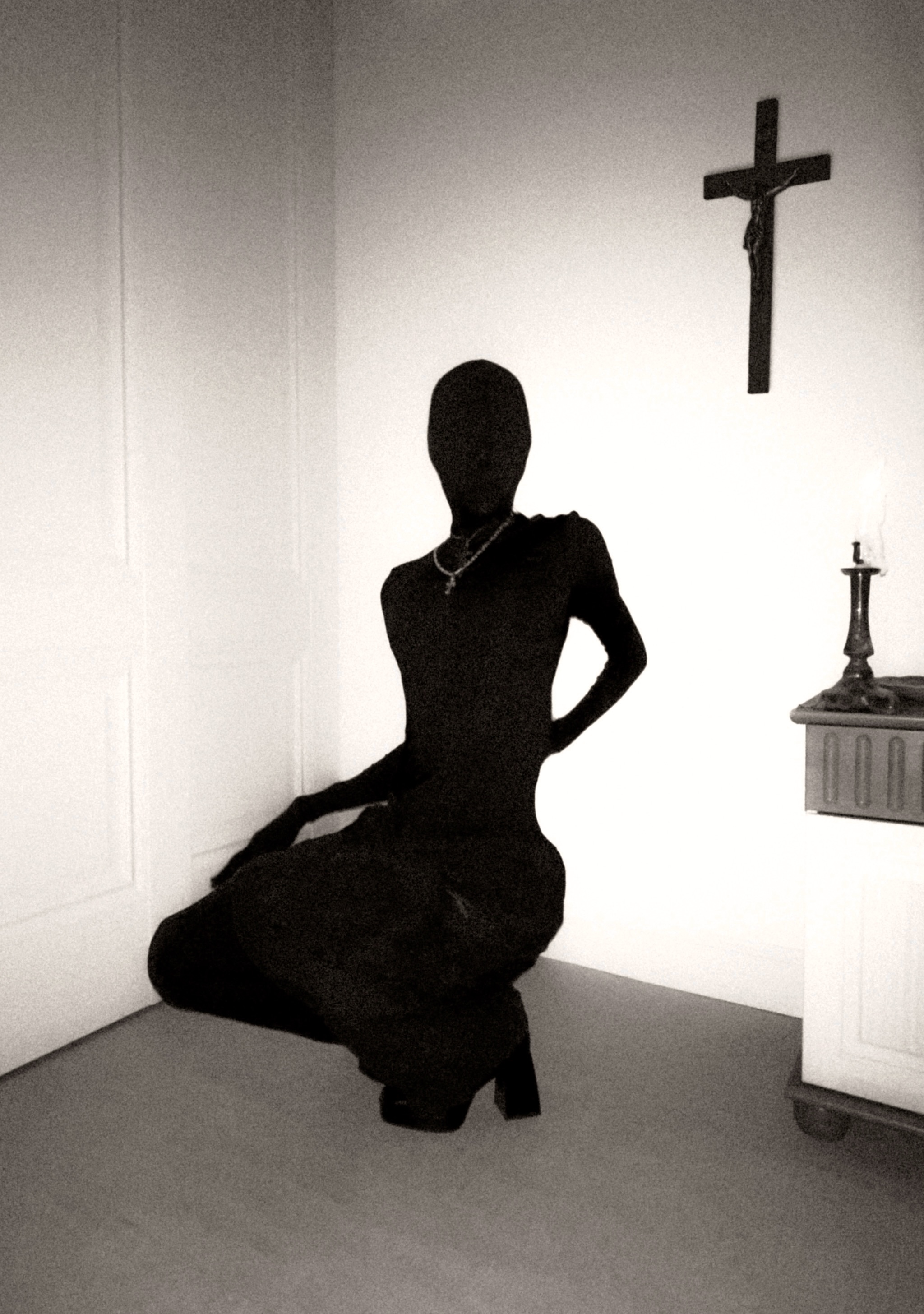 Spread from TheHunterIssue Vol.1 by Leo Shmunk, faceless figure in a black full-body morphsuit kneeling in a white-walled corner beneath a wooden crucifix, candle and small altar at right, 2026. Photo: Leo Shmunk.