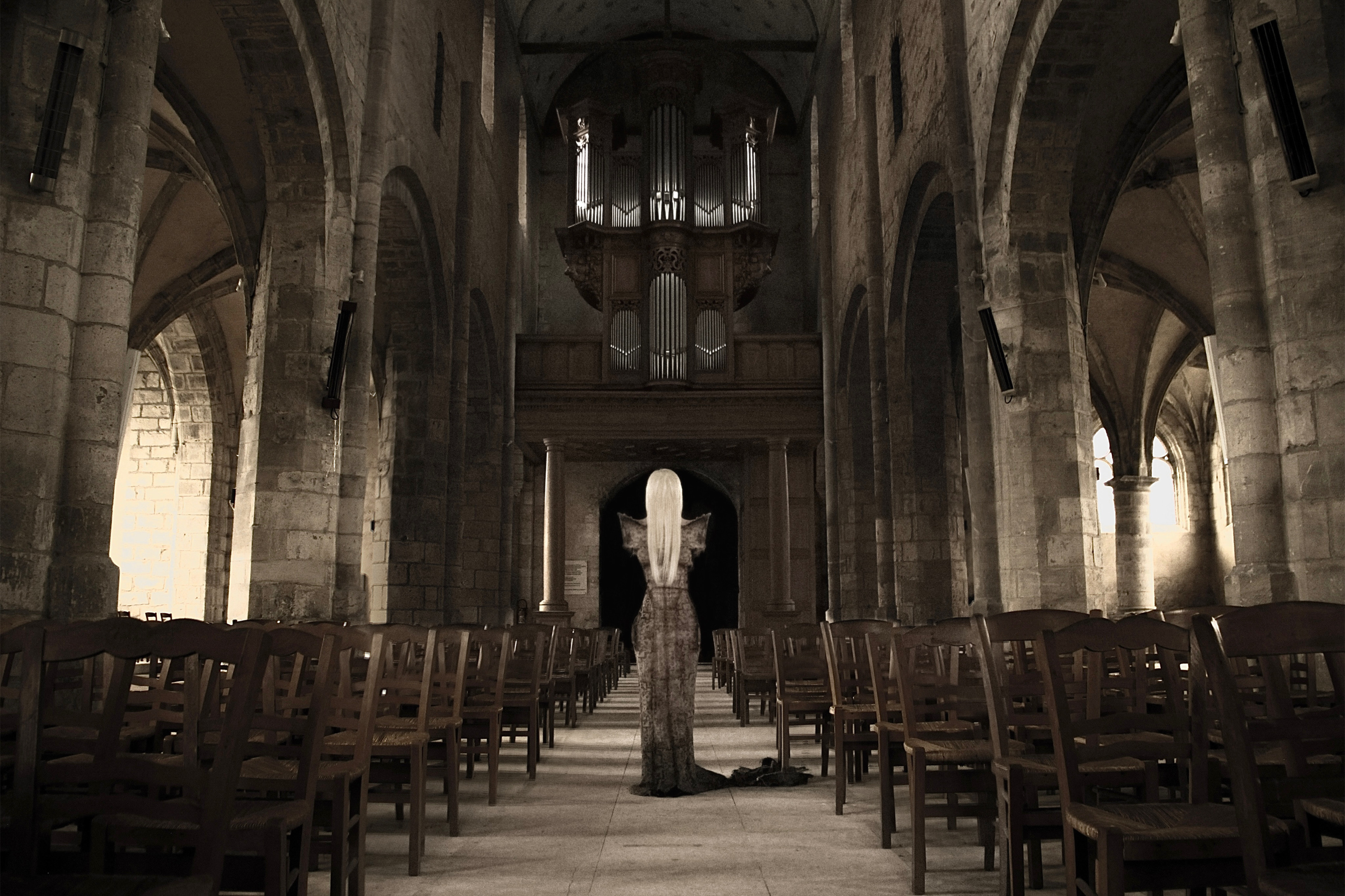Spread from TheHunterIssue Vol.1 by Leo Shmunk, sepia nave interior with a pale-haired figure in a long gown standing alone at the center aisle beneath an organ loft, rows of empty wooden chairs flanking, 2026. Photo: Leo Shmunk.