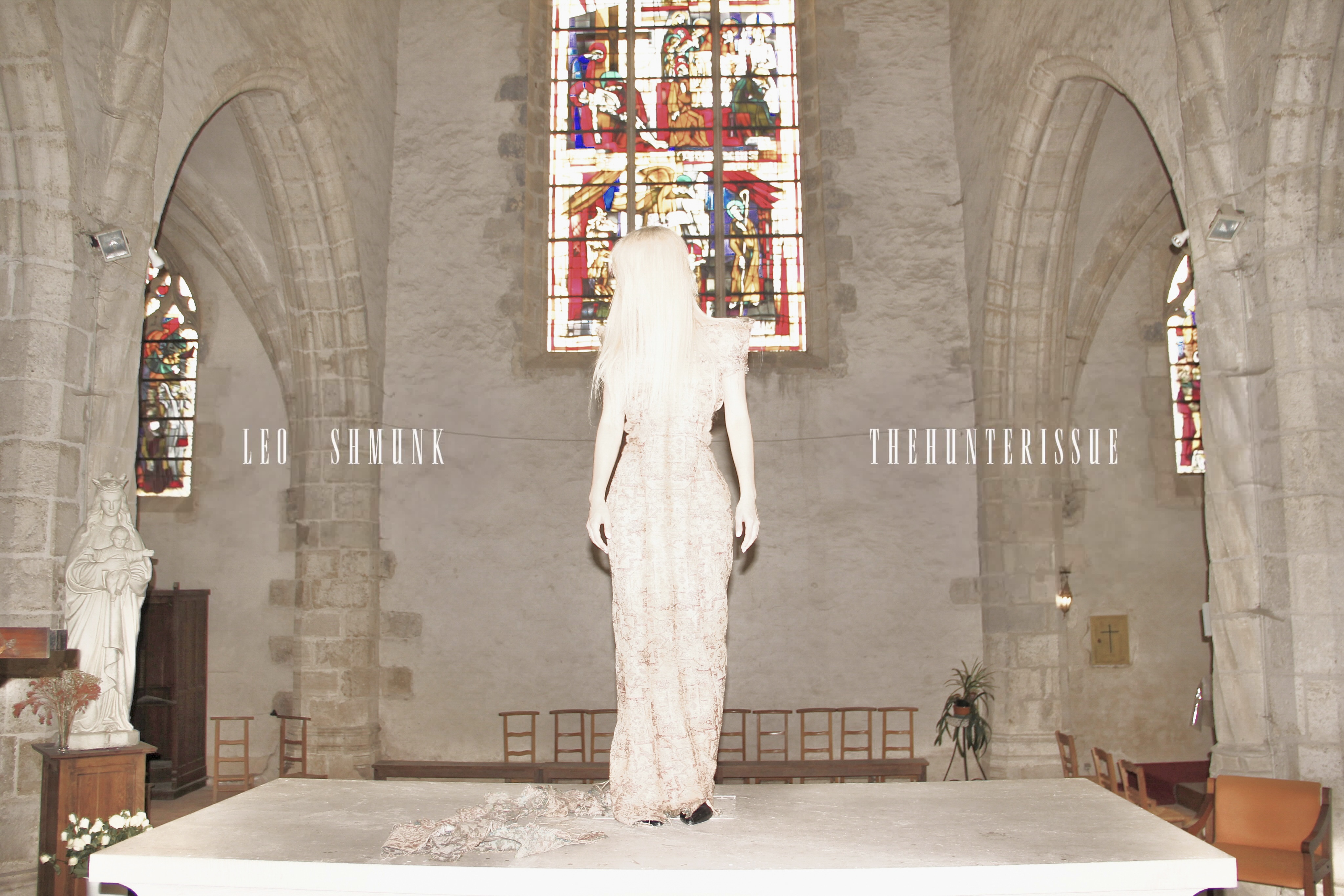 Spread from TheHunterIssue Vol.1 by Leo Shmunk, white-haired figure in a pale lace gown facing a stained-glass window inside a vaulted stone church, LEO SHMUNK and THEHUNTERISSUE wordmarks flanking the nave, 2026. Photo: Leo Shmunk.