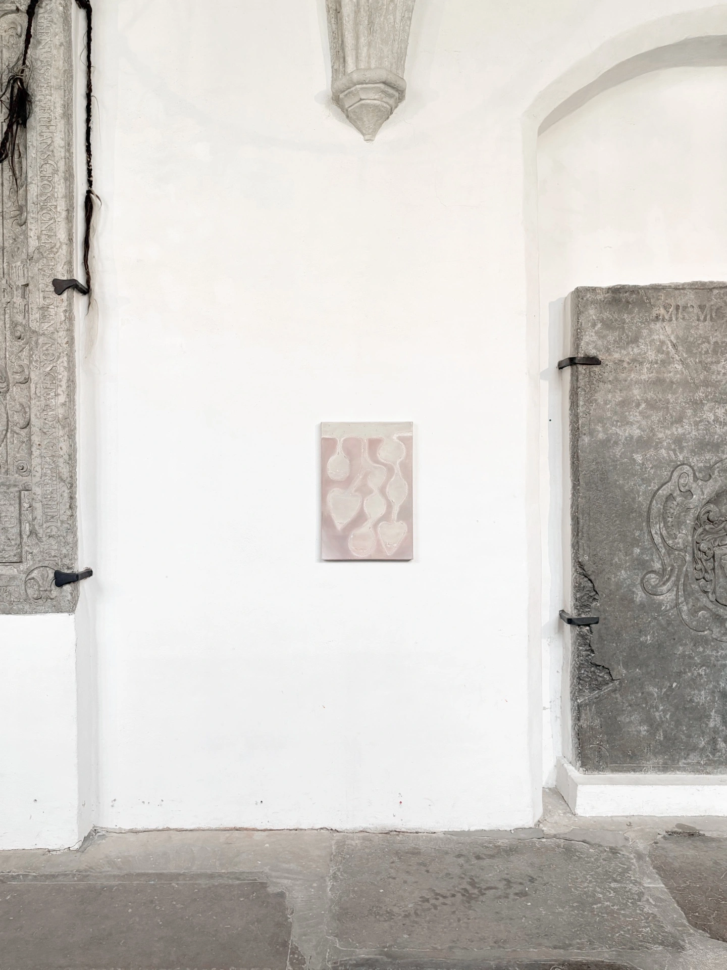 Angelika Puff, Thaw, a small rose-toned canvas of pendulous seed-like forms mounted on a white wall between a carved stone epitaph and a grey medieval tomb slab.
