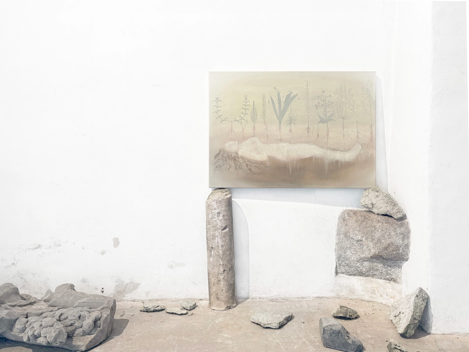 Angelika Puff, Cycle, a small horizontal painting of rooted plant specimens above a reclining pale figure, propped on rough stone plinths amid scattered rocks on the church floor.