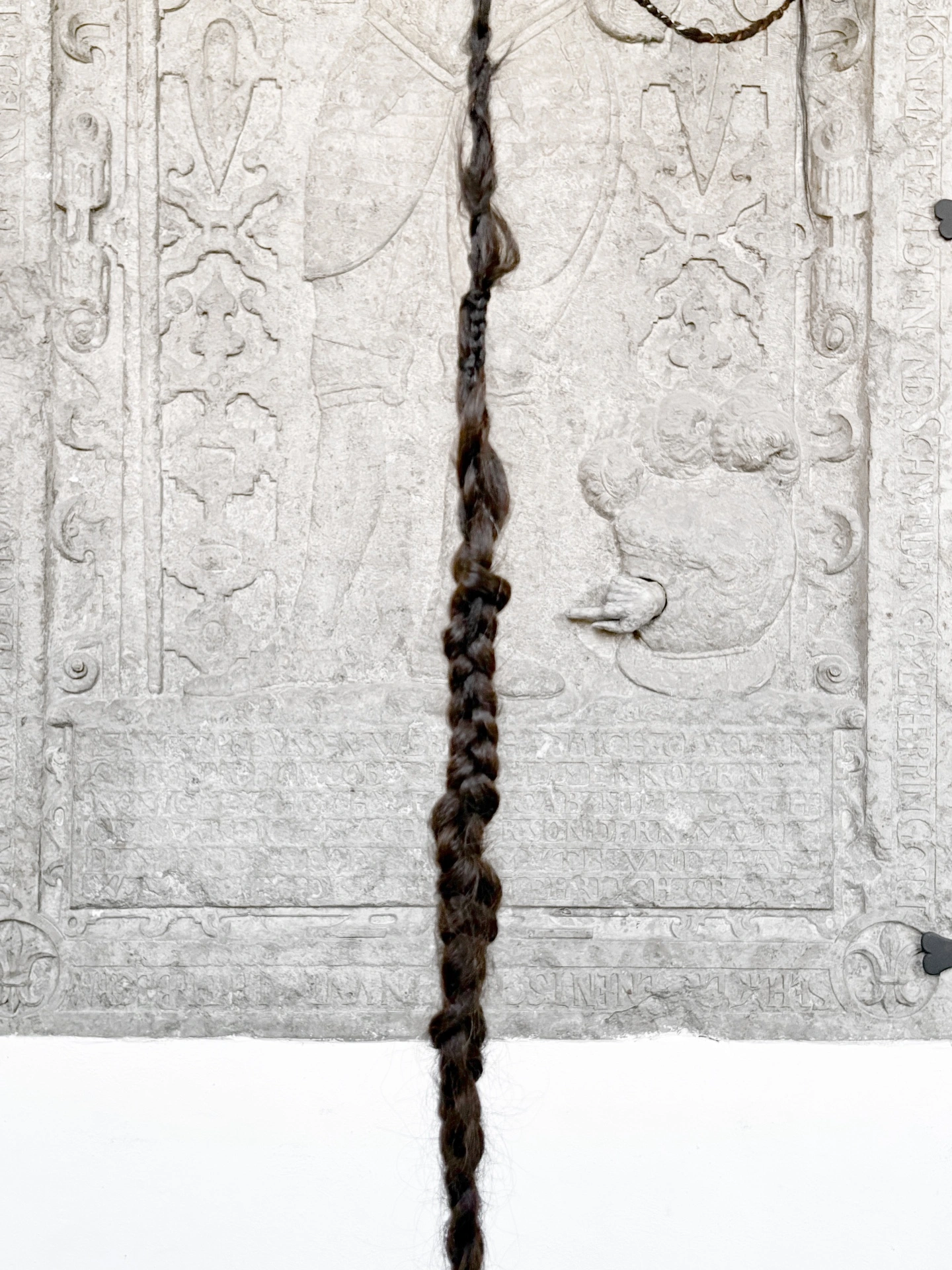 Angelika Puff, Bird Cage detail, 2025 — single long braid of human hair running down the face of a medieval Latin inscription stone at Galeria EL, hair and stone in contact.