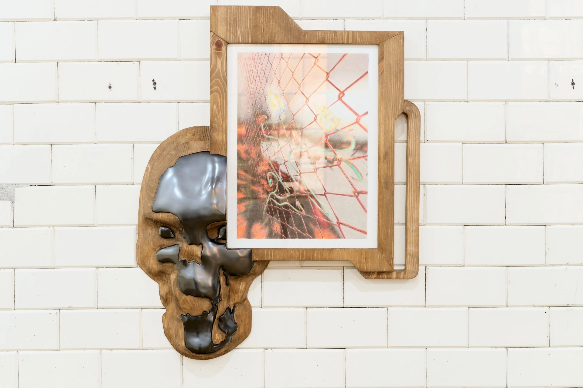 Tristan Gac, chrome half-skull cast into carved pine gripping a drawing of orange flame behind chain-link fence with green tendrils