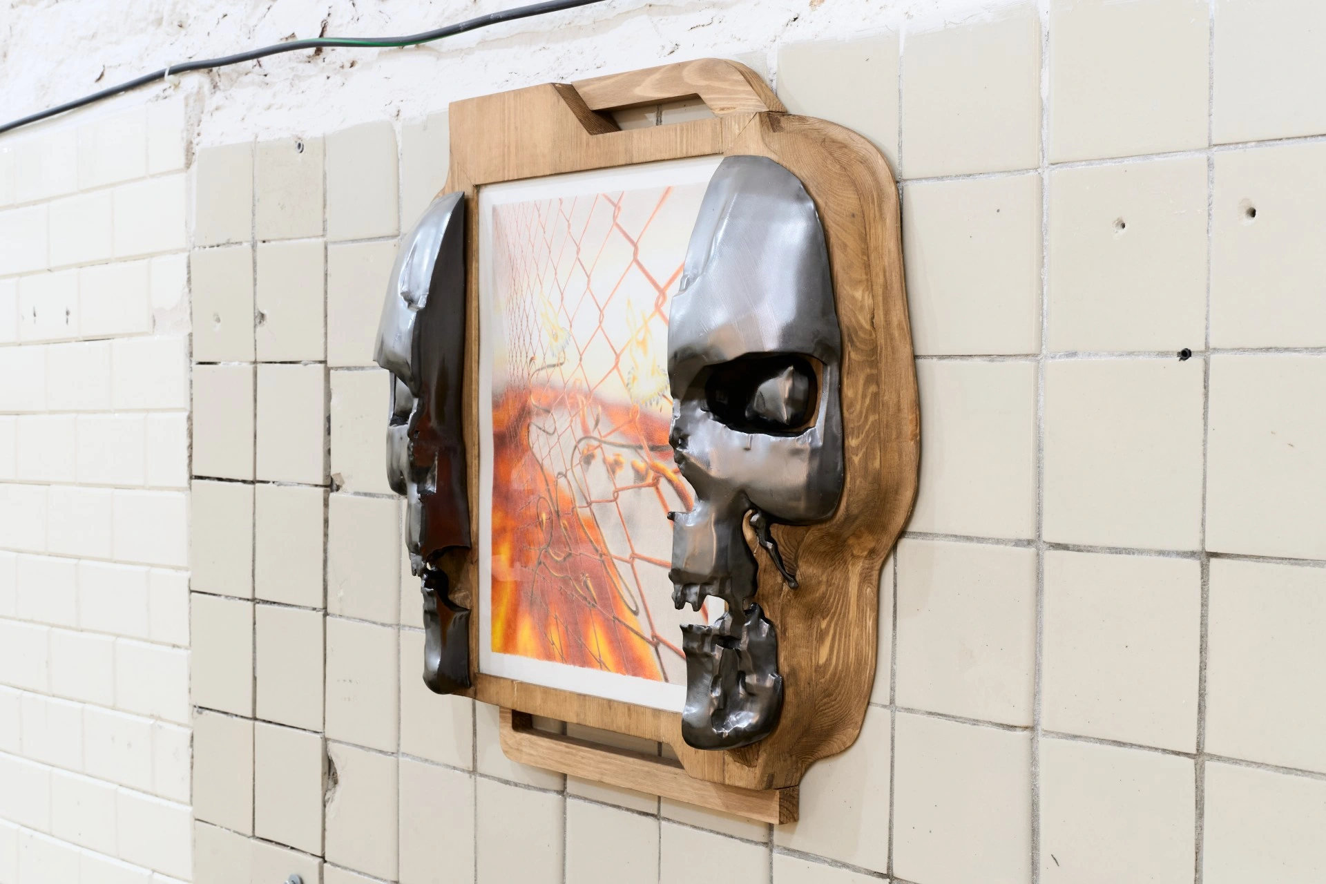 Tristan Gac, angled view of a gunmetal half-skull cast into pine gripping a framed drawing of orange flame behind chain-link fence