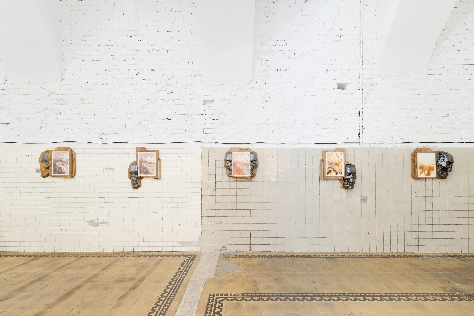 Chrome Terminator skulls inlaid into carved pine boards grip framed pencil drawings of chain-link fences dissolving into flame and flowers rising from bone-strewn rubble, installed on white tile and vaulted brick.