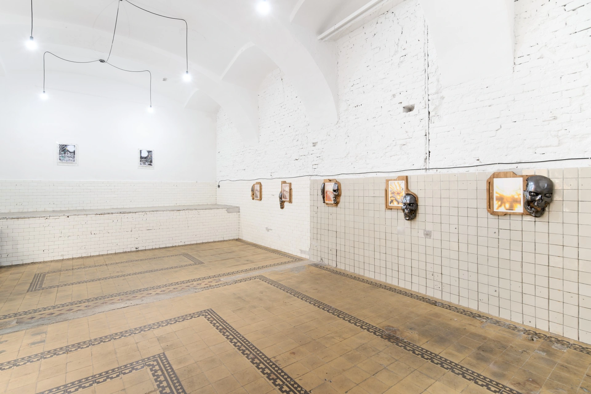 Tristan Gac, installation view along white tiled wall with vaulted brick, a row of chrome skulls on carved pine boards gripping framed drawings, encaustic tile floor