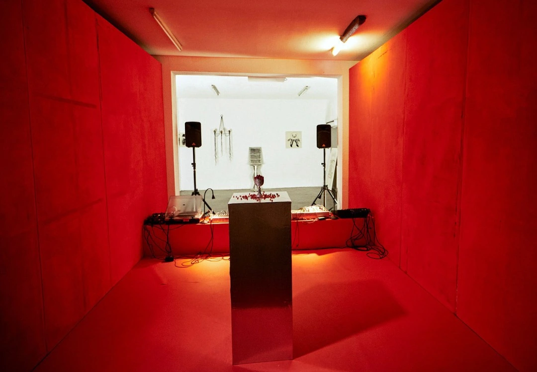 Performance set, Seeds know no tree., New Fears Gallery, Berlin, 2026: deep red-draped chamber with modular synthesizers on flanking tables, concrete plinth centre, PA speakers on tripods, doorway opening to white gallery beyond. Photo: Sandro Mosco