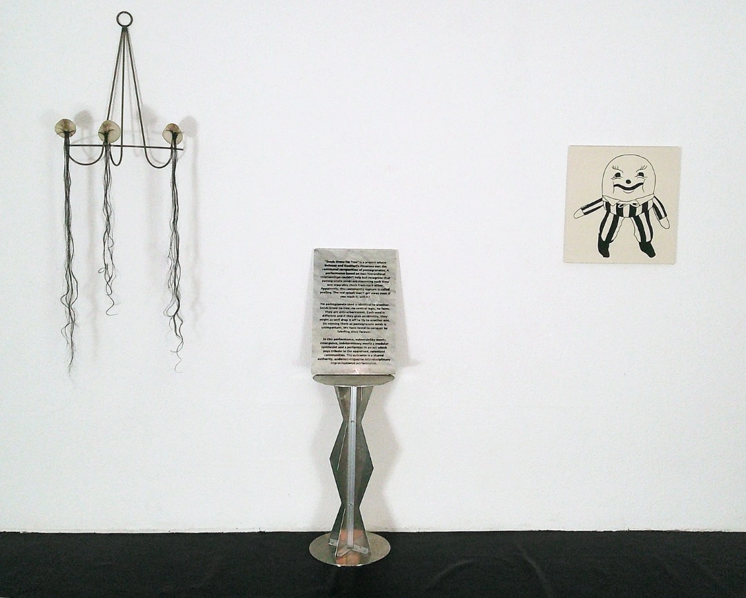 Installation view, Seeds know no tree., New Fears Gallery, 2026: metal chandelier-like sculpture with hanging hair filaments, steel stele with engraved curatorial text on plinth, naive cartoon painting on white gallery wall. Photo: Sandro Mosco