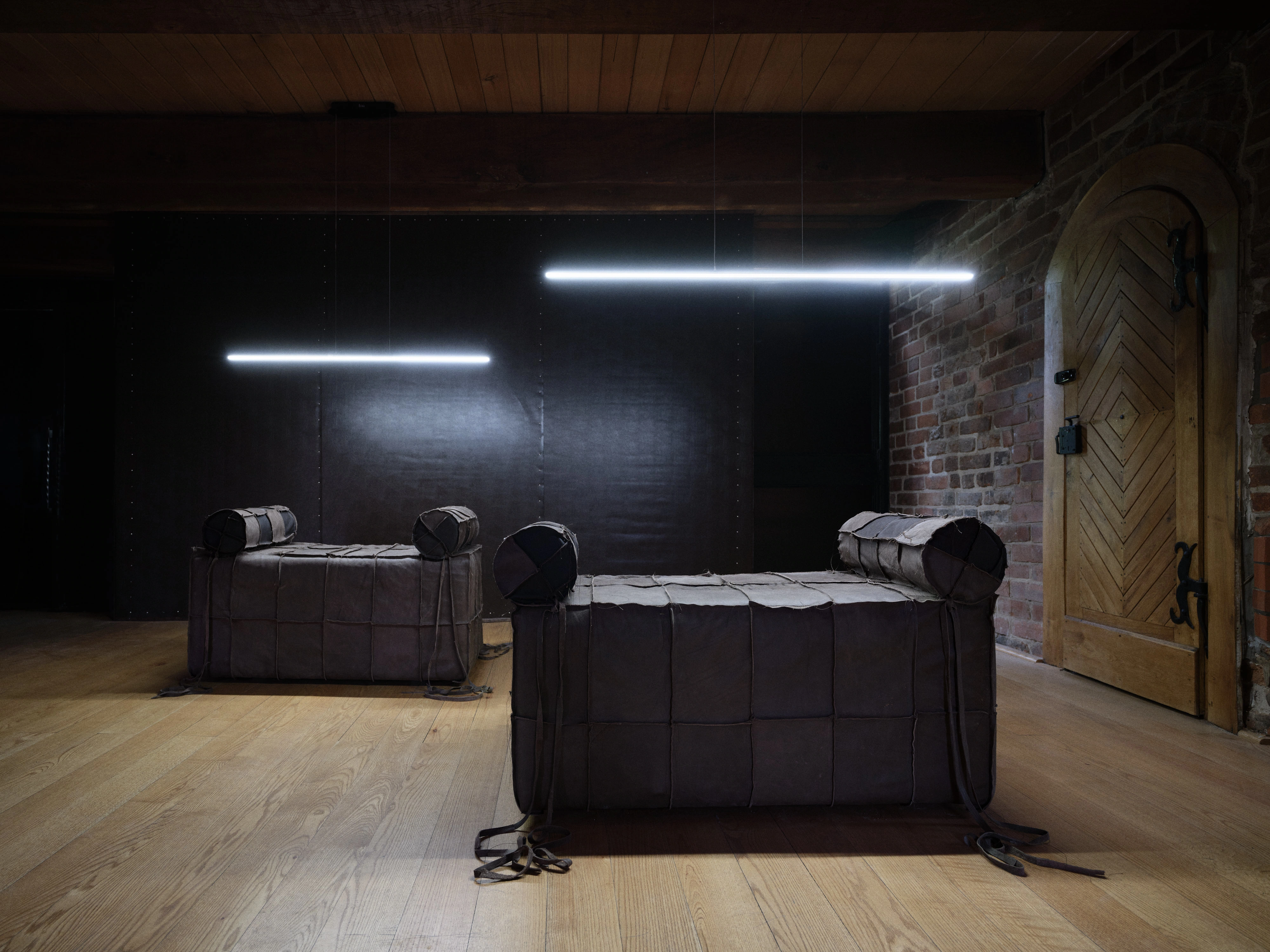 Jan Baszak, two Catafalque biers upholstered in patched brown leather with bolsters at each end, ties trailing across pale floorboards beside an arched timber door