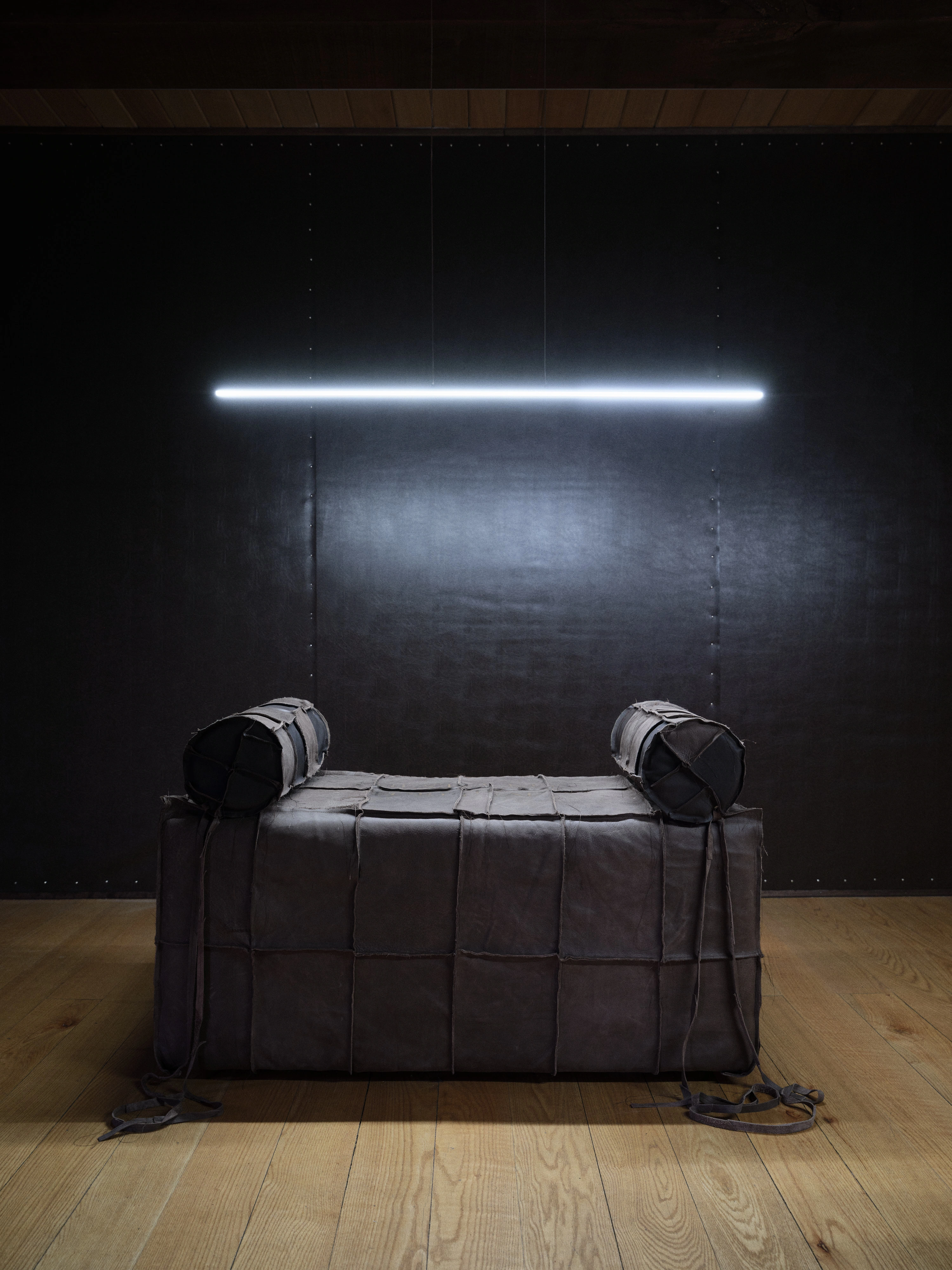 Jan Baszak, a single Catafalque centred beneath a suspended tube of light against a black riveted wall panel, leather ties pooled on the floor