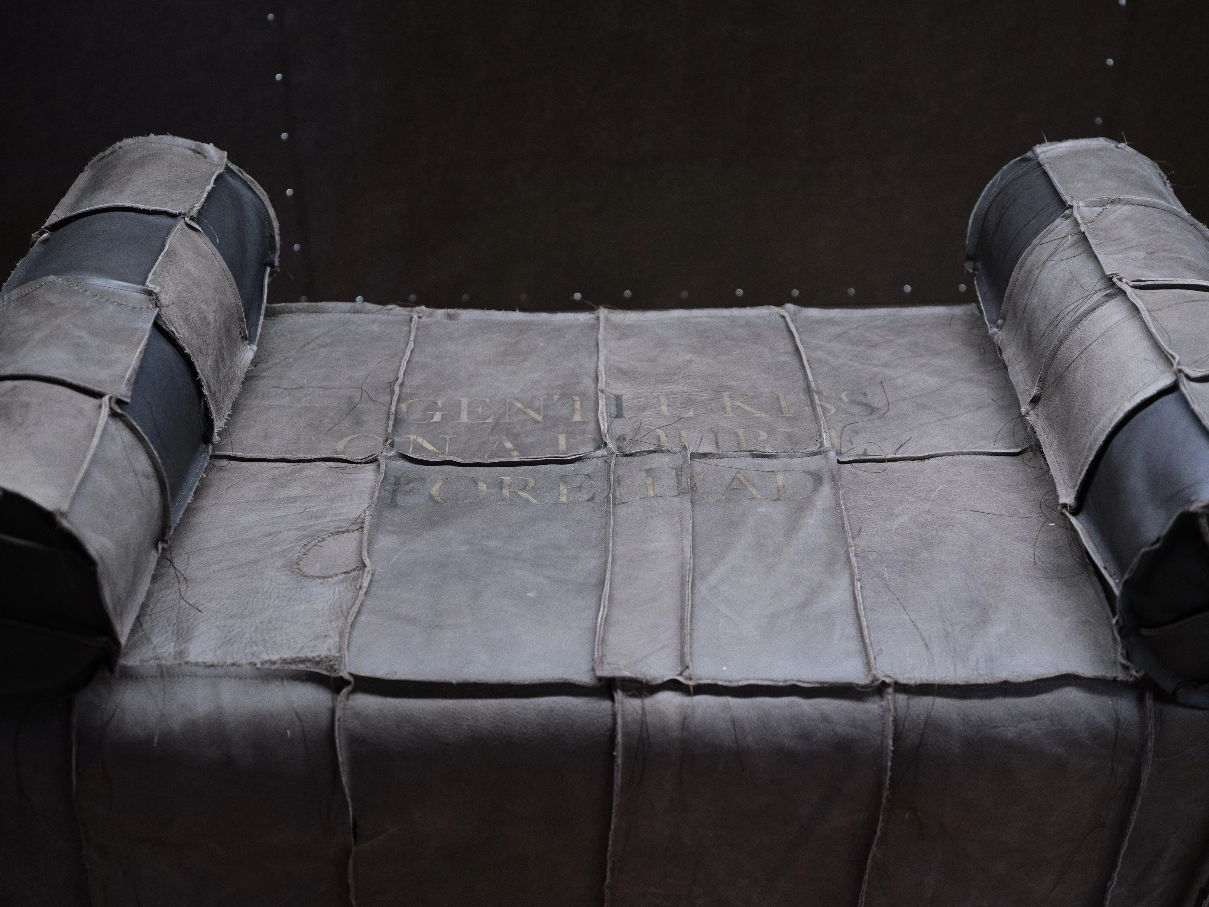 Jan Baszak, the engraved top panel of a Catafalque, the words of the exhibition title burned faintly into grey leather between two bolsters