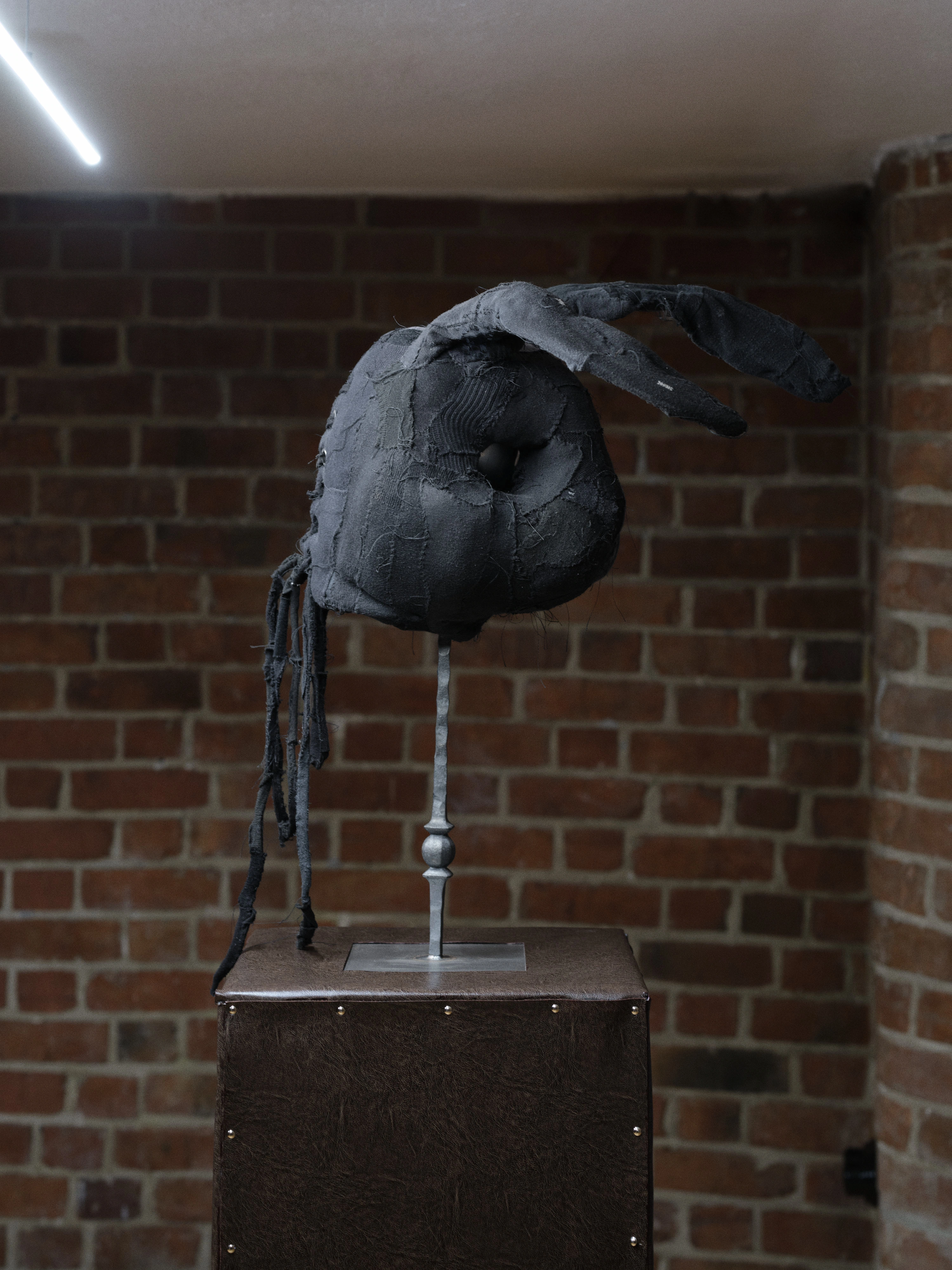 Jan Baszak, Rabbit Mask on a metal stand with one ear folded upright and one laid back, strips of black fabric hanging beside the plinth