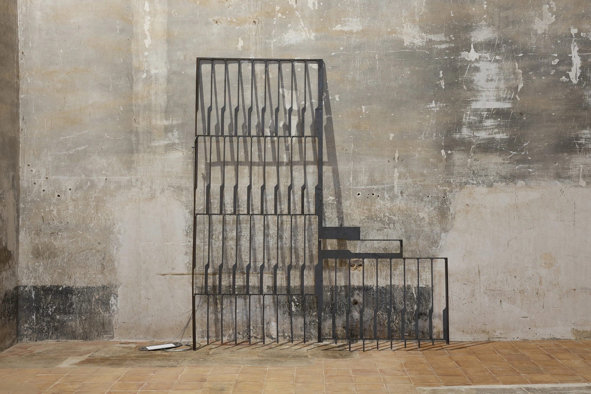 Jordi Mitjà's stepped wrought-iron railing leaning against a bare plaster wall, a blank paper label and steel cable on the tiles beside it.