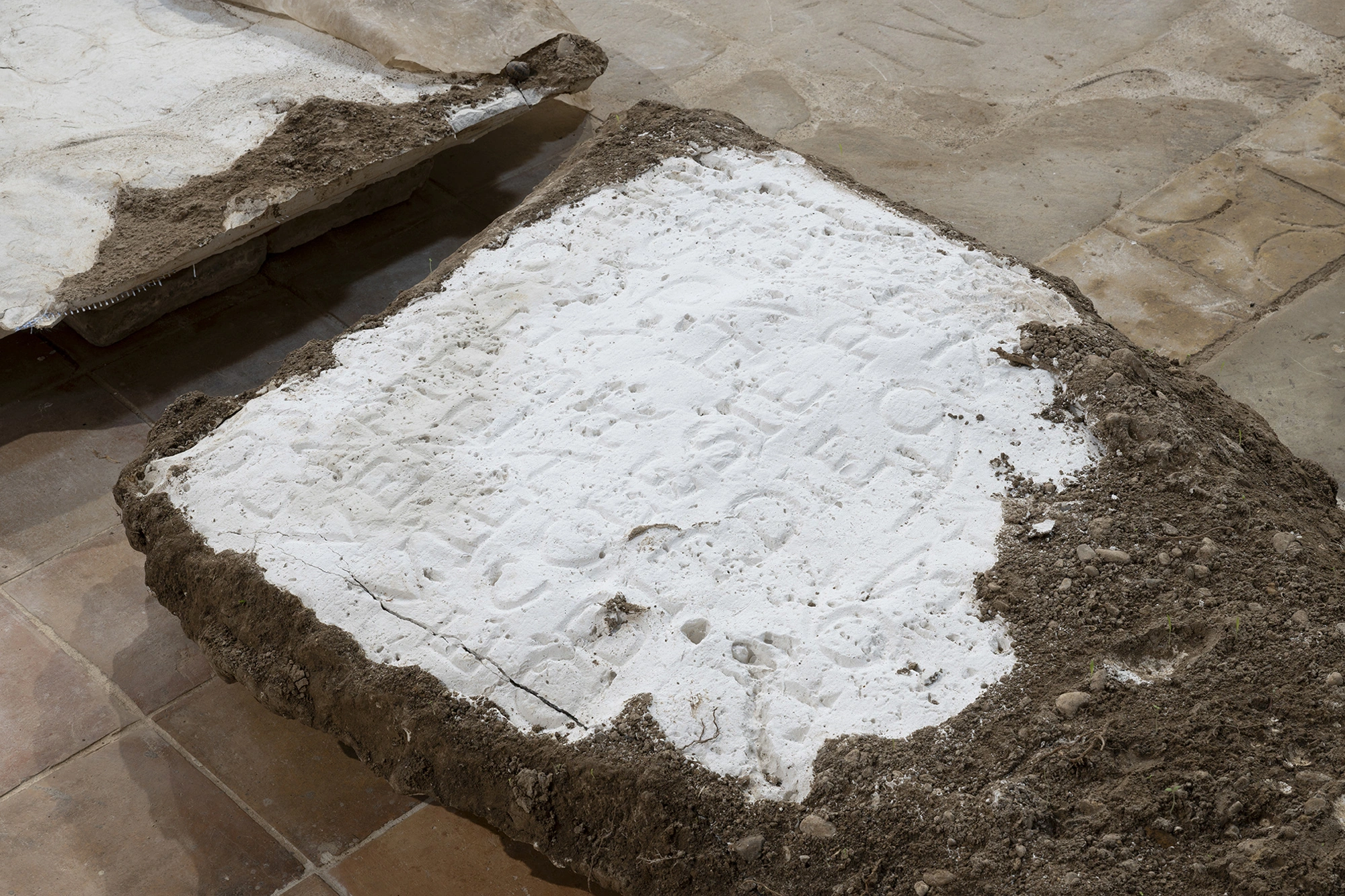 Detail of a plaster cast from Monia Ben Hamouda's Ghost series, its inscription standing in reversed relief, the slab rimmed with dark soil.