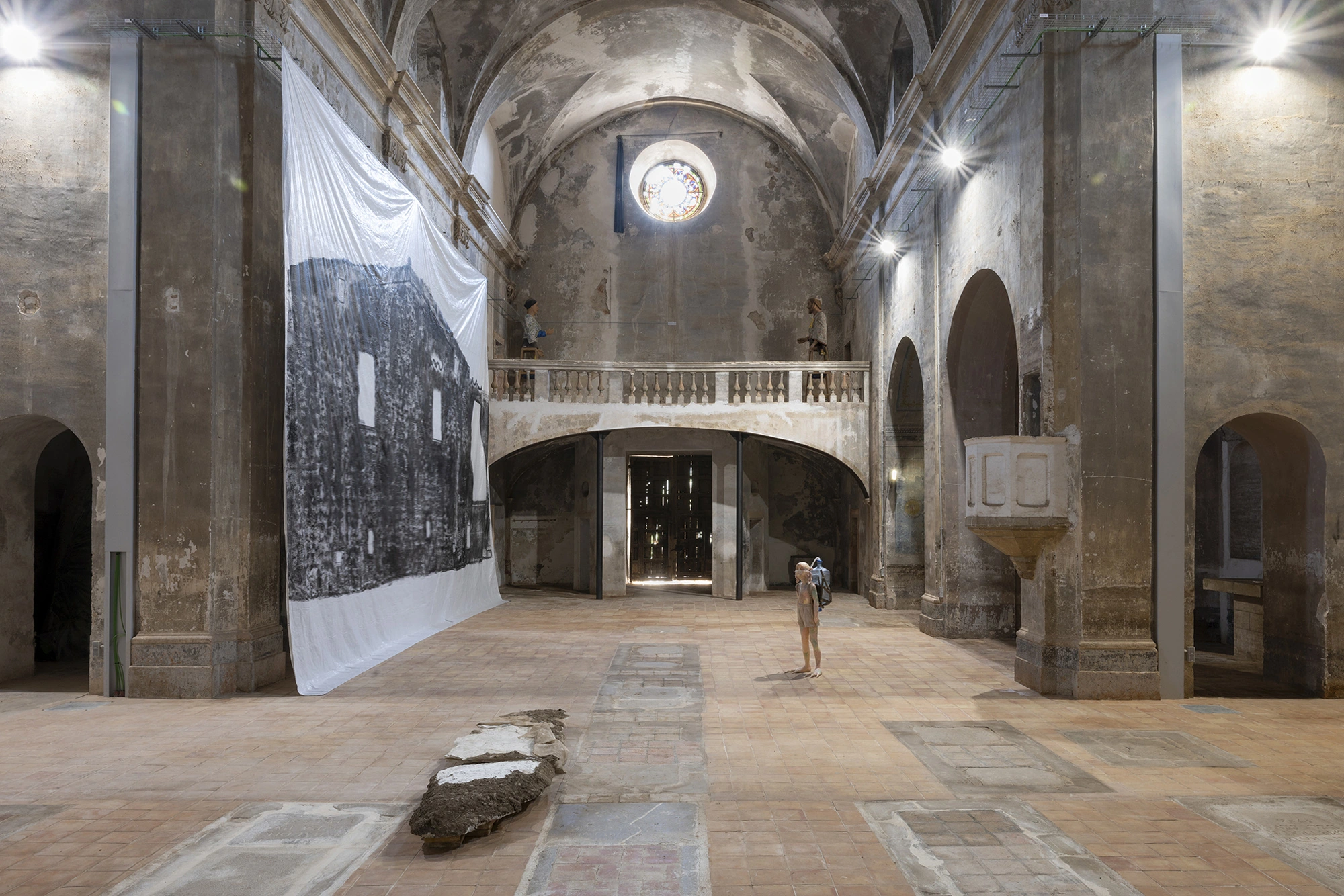 The nave of the Church of Sant Feliu, Lladó, with Andrew Birk's Floating House hanging from the cornice, Michele Gabriele's July 2nd standing alone on the tiles and Monia Ben Hamouda's Ghost works in the foreground.