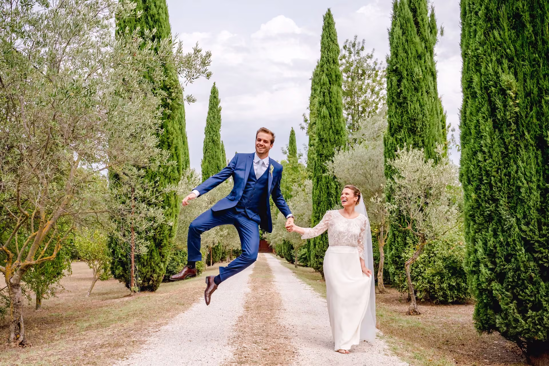 Un couple de mariés souriants marchant sur une allée bordée de cyprès, le marié en costume bleu sautant joyeusement.
