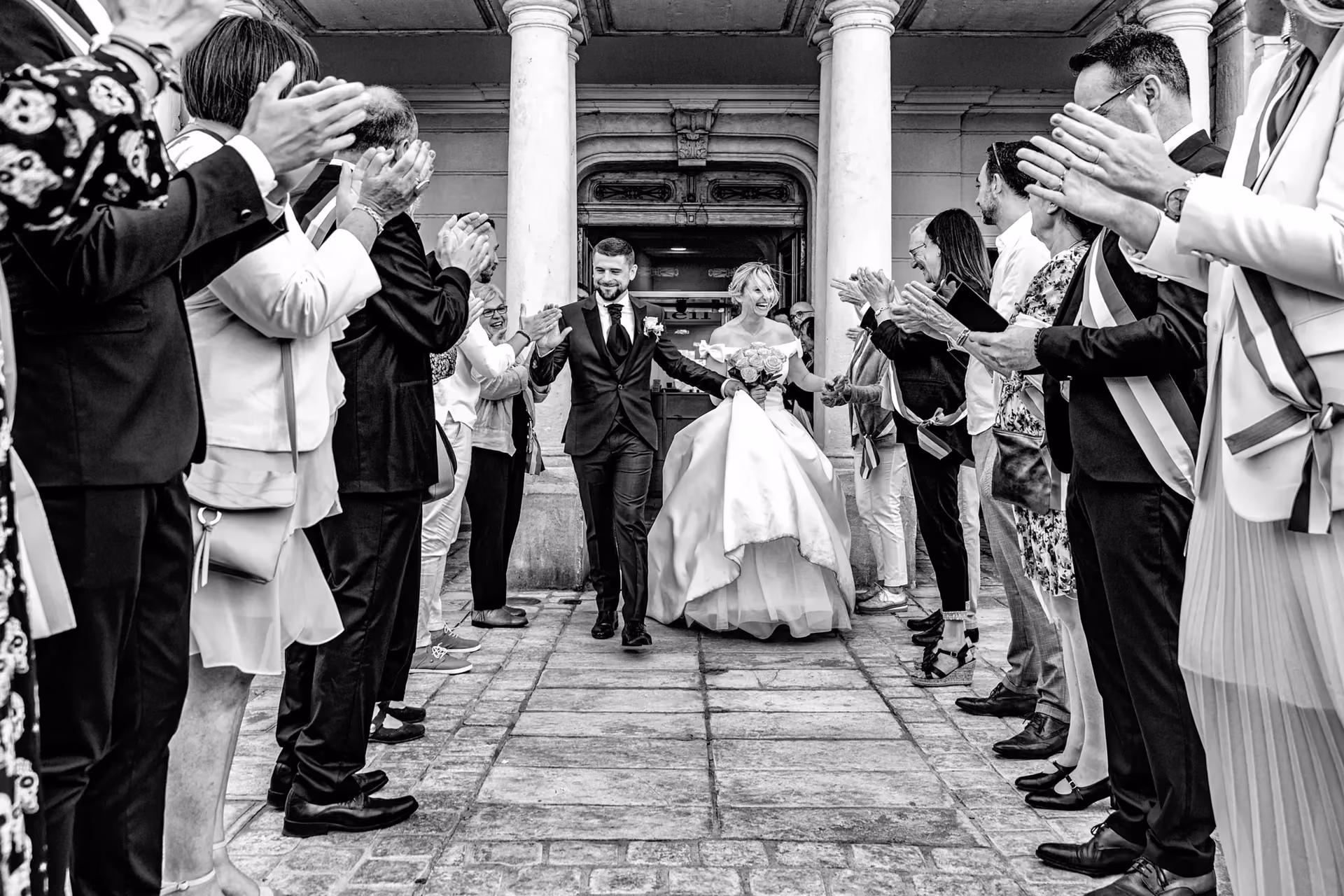 Couple de mariés souriants quittant un bâtiment historique sous un applaudissement chaleureux.