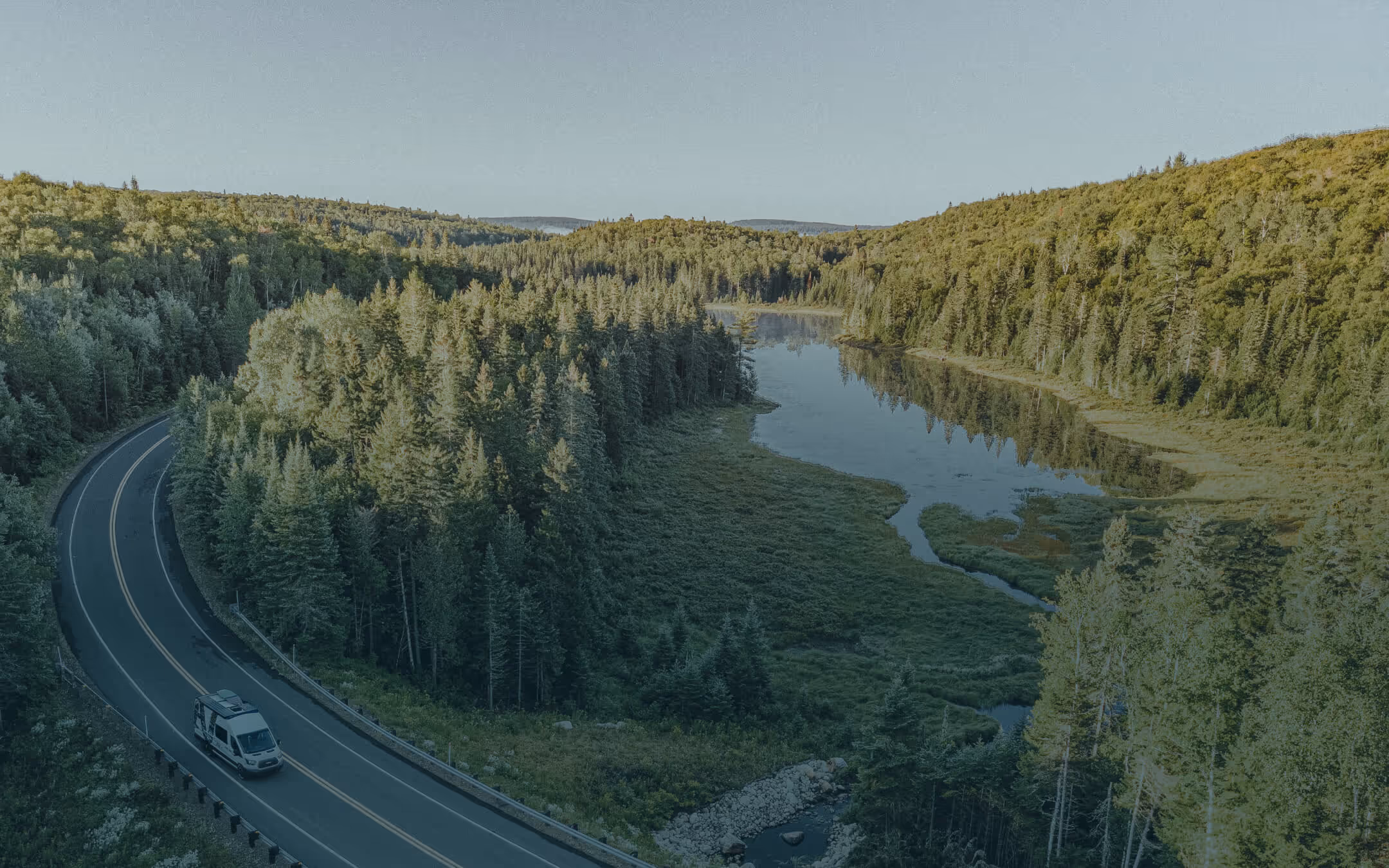 A white camper van driving on a winding road through dense green forest alongside a calm river under clear sky.