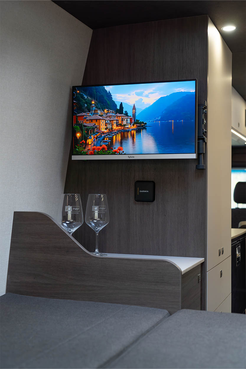 Interior view of a modern living space with a wall-mounted flat-screen TV displaying a scenic lakeside village at dusk, two empty wine glasses on a wooden ledge, and a Garmin device mounted below the TV.