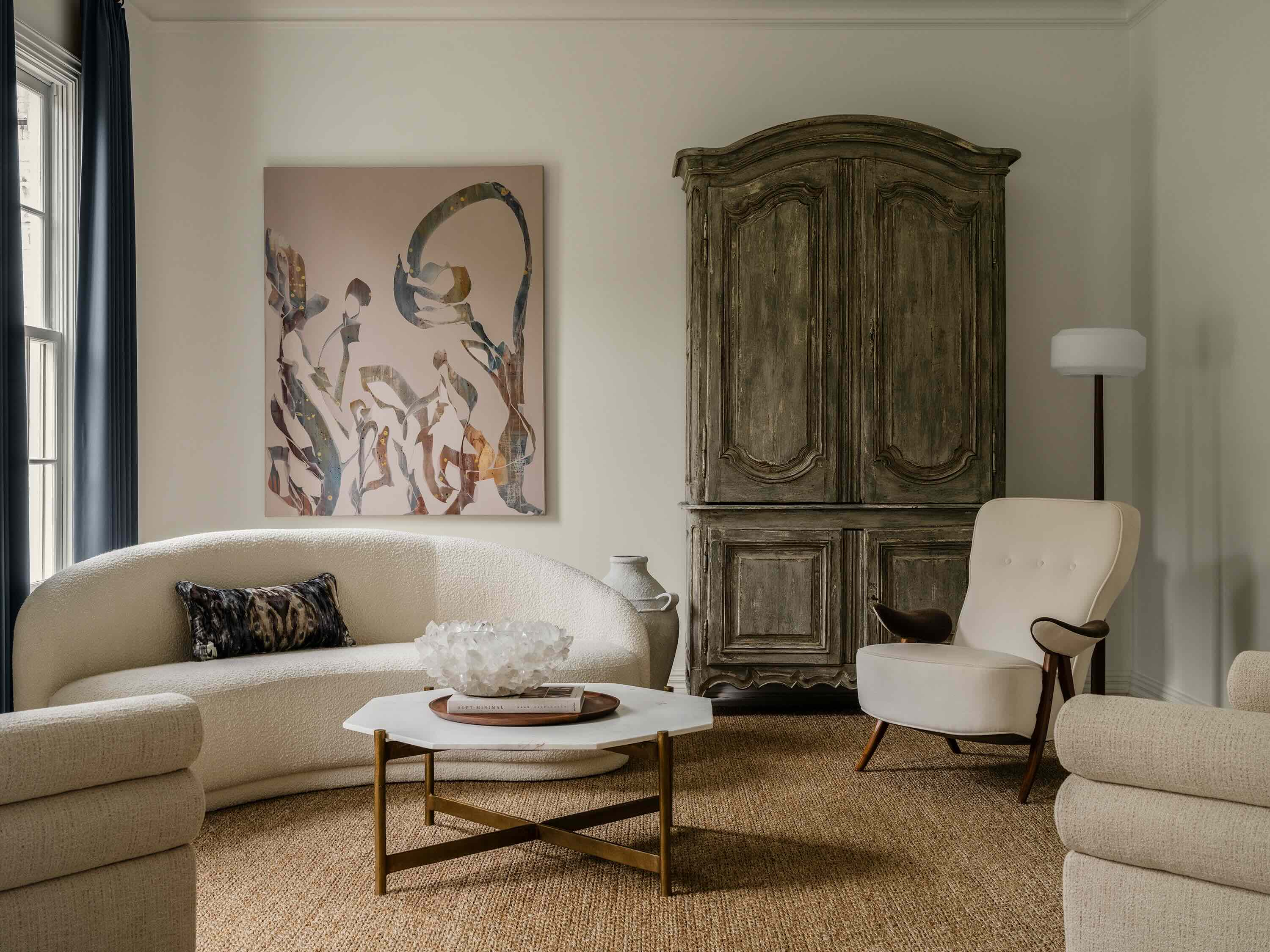Living room with curved beige sofa, abstract wall art, antique wooden cabinet, cream armchair, hexagonal marble coffee table, and floor lamp.